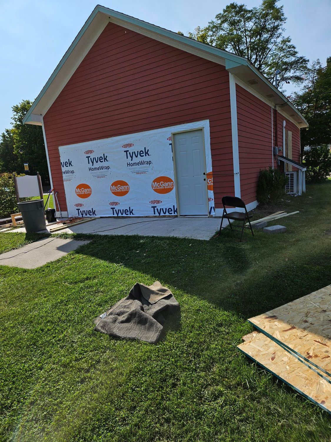 Red house under construction with white Tyvek wrap over the garage door and lawn tools in front