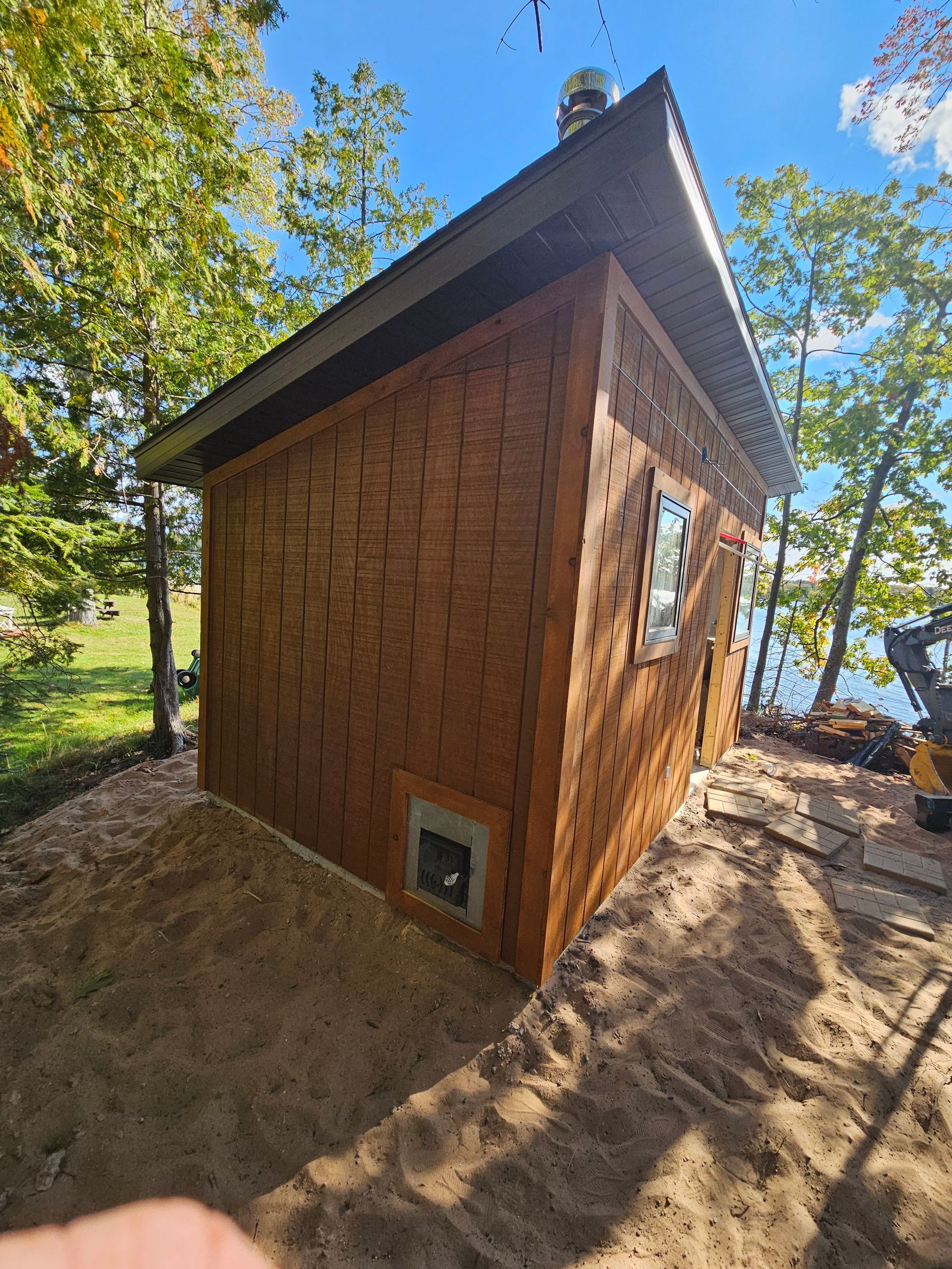 Small wooden cabin with a sloped roof on a wooded lakeshore, sunlight and trees around it.