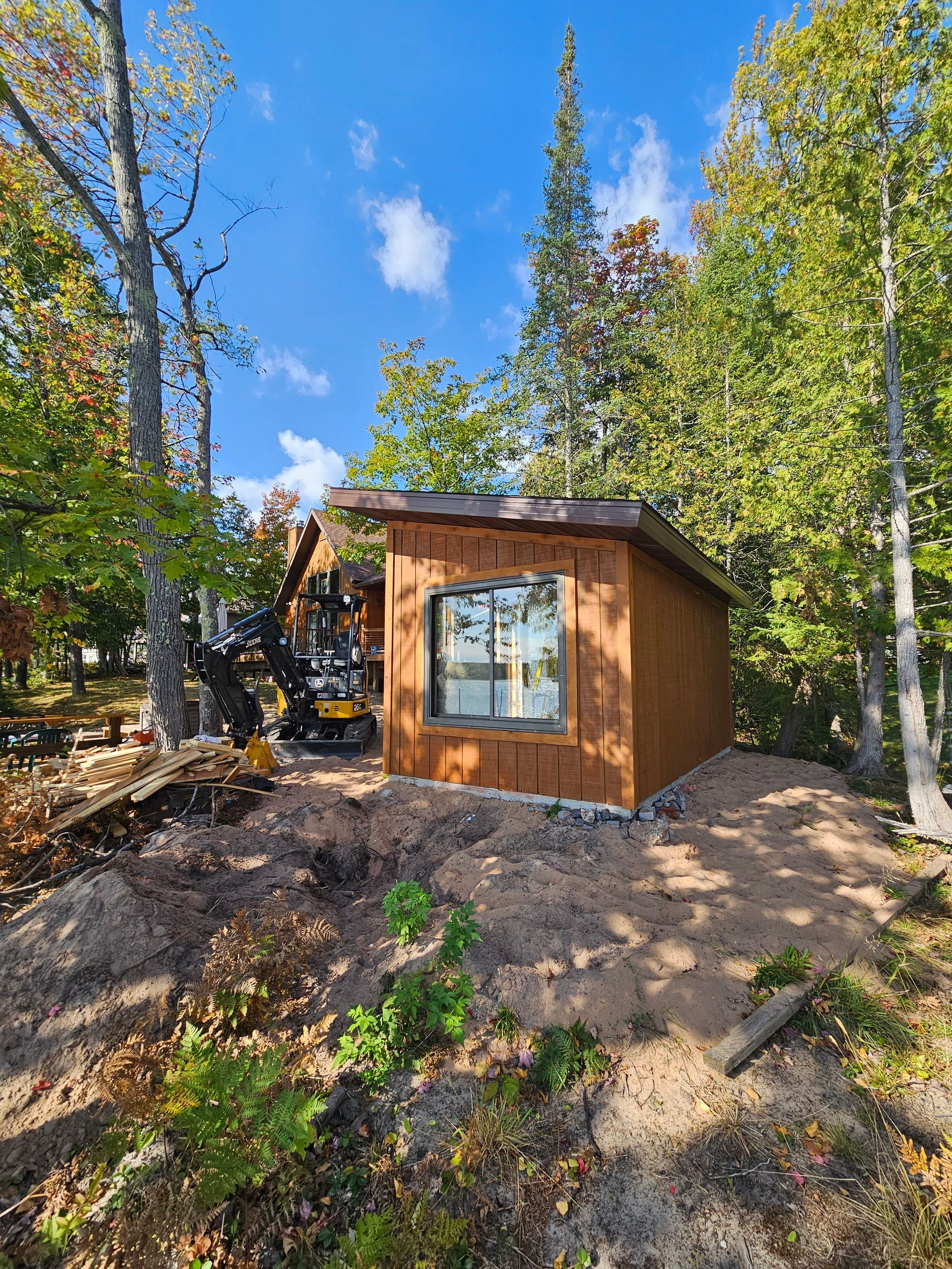 Small wooden cabin with a window in a sunny forest clearing, surrounded by tall trees and blue sky.
