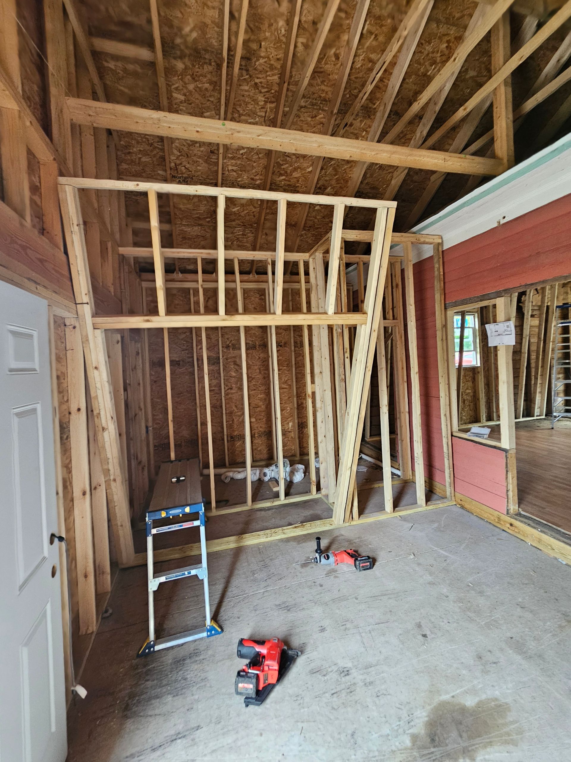Partially framed interior room with exposed roof trusses, tools on the floor, and a doorway to a red exterior wall
