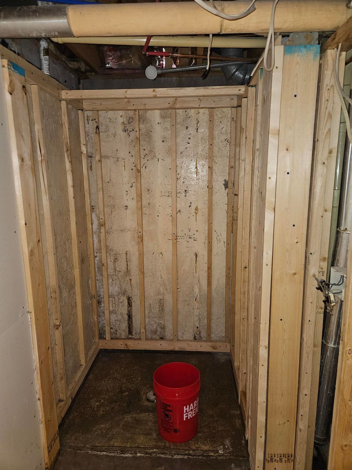 Old wooden basement alcove with exposed pipes and a red bucket on the floor