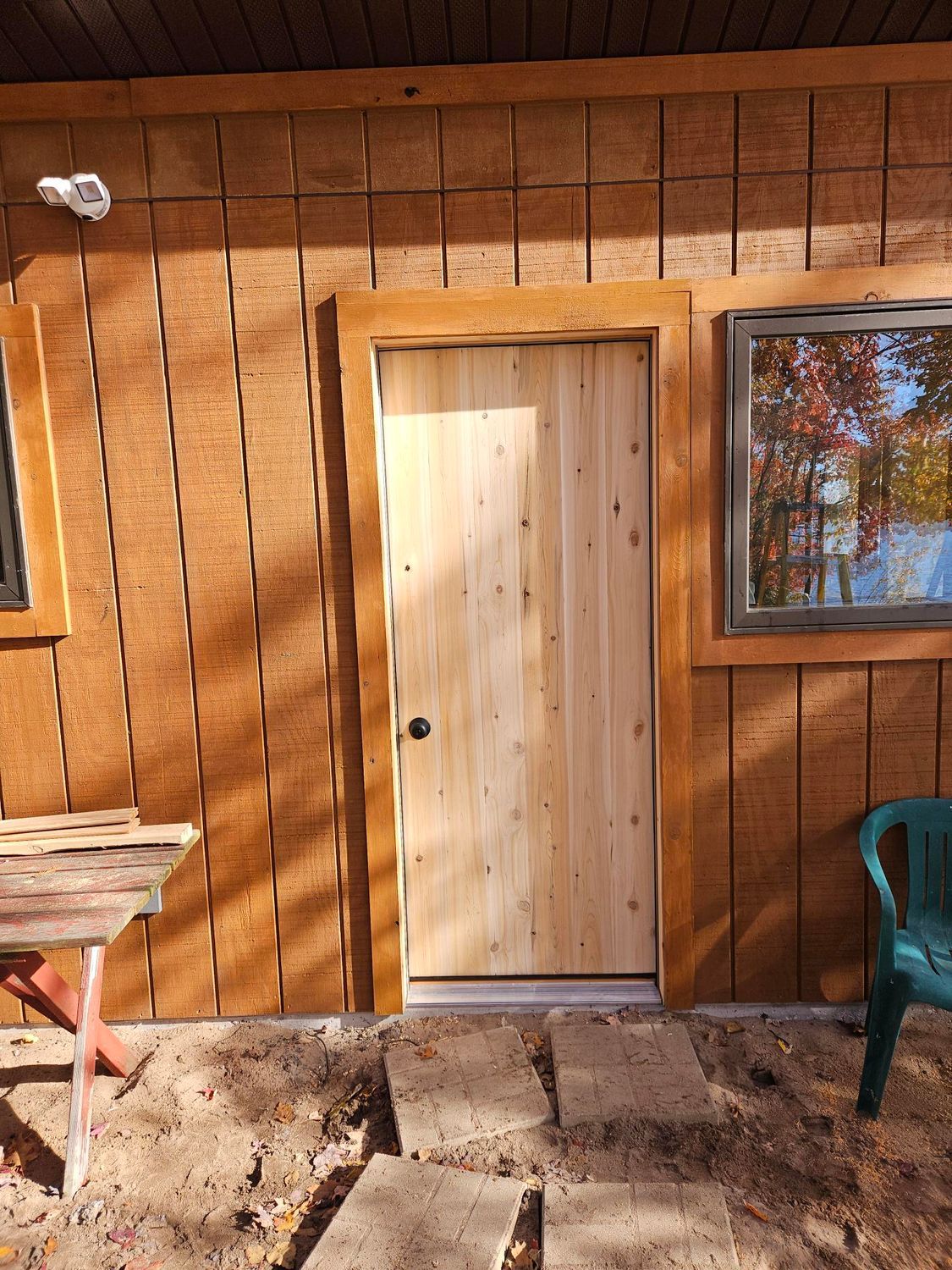 Wooden cabin exterior with a beige door and window, patio stones, and a green chair in sunlight