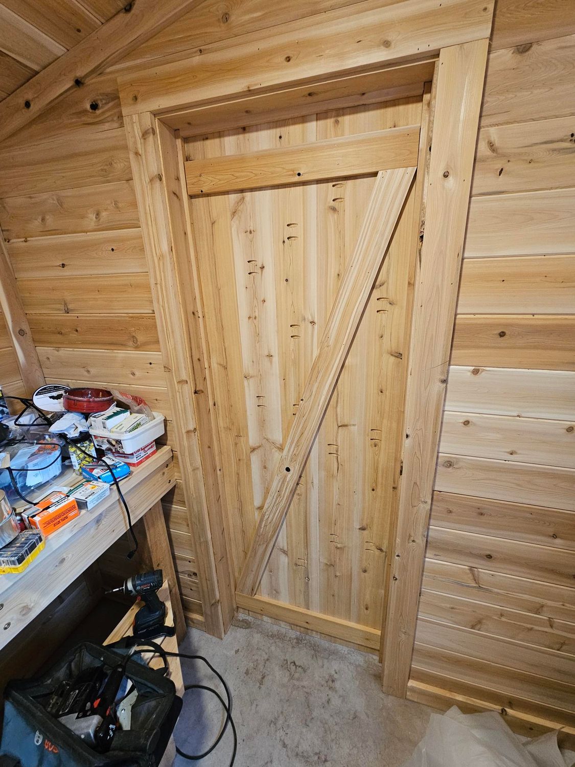 Wood-paneled room with a small wooden door; cluttered workbench at left and concrete floor.