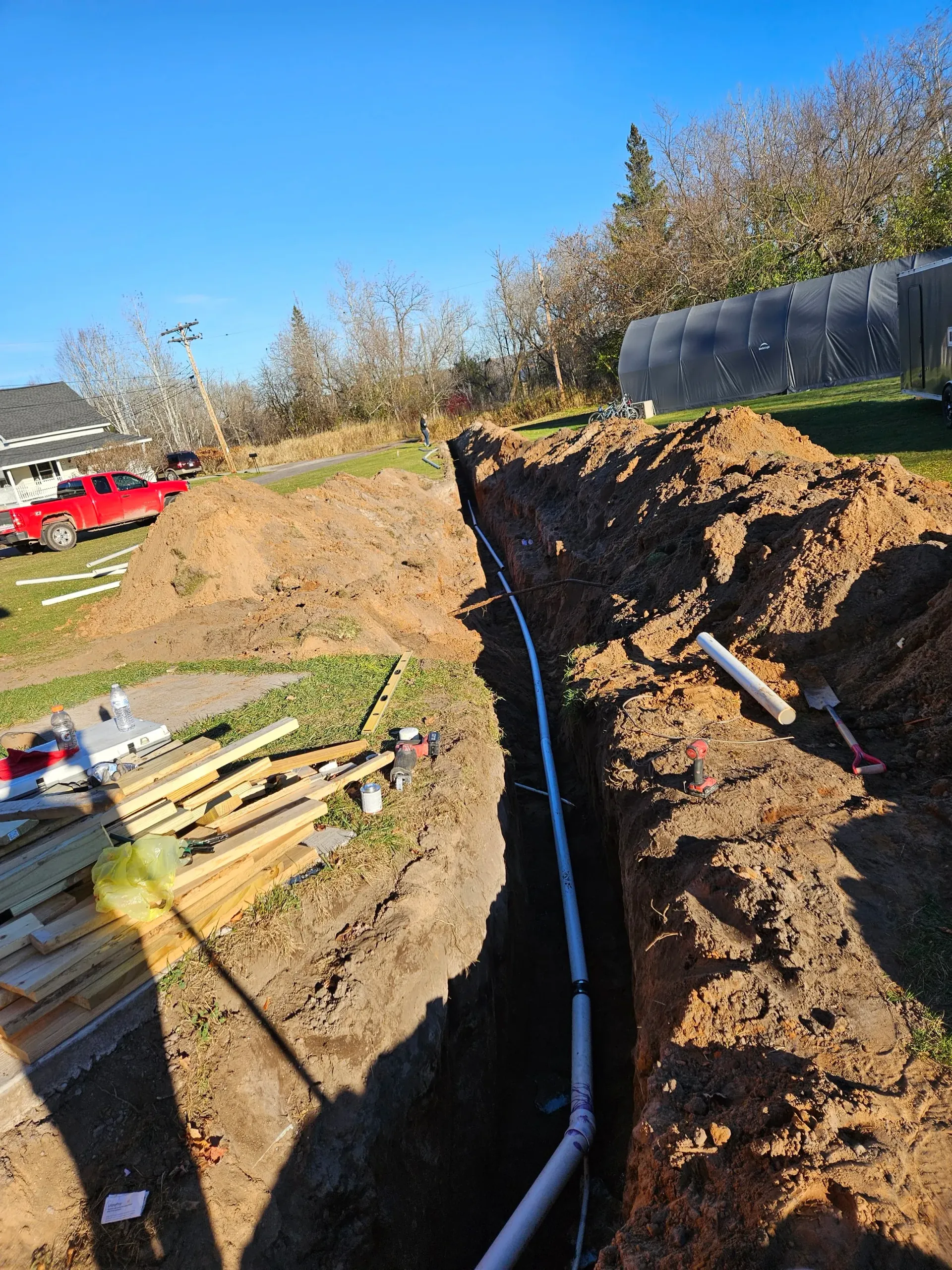 Trench excavation with exposed black pipe, dirt piles, tools, and a red truck in the background outdoors