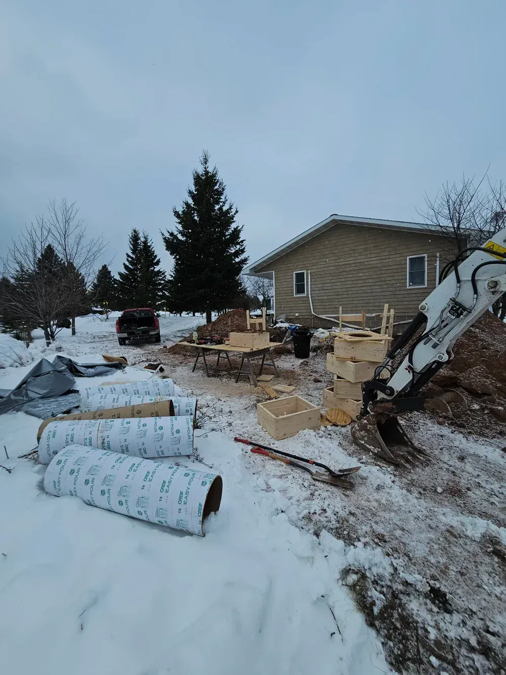 Snow-covered yard with construction debris, pipes, and a house under overcast sky