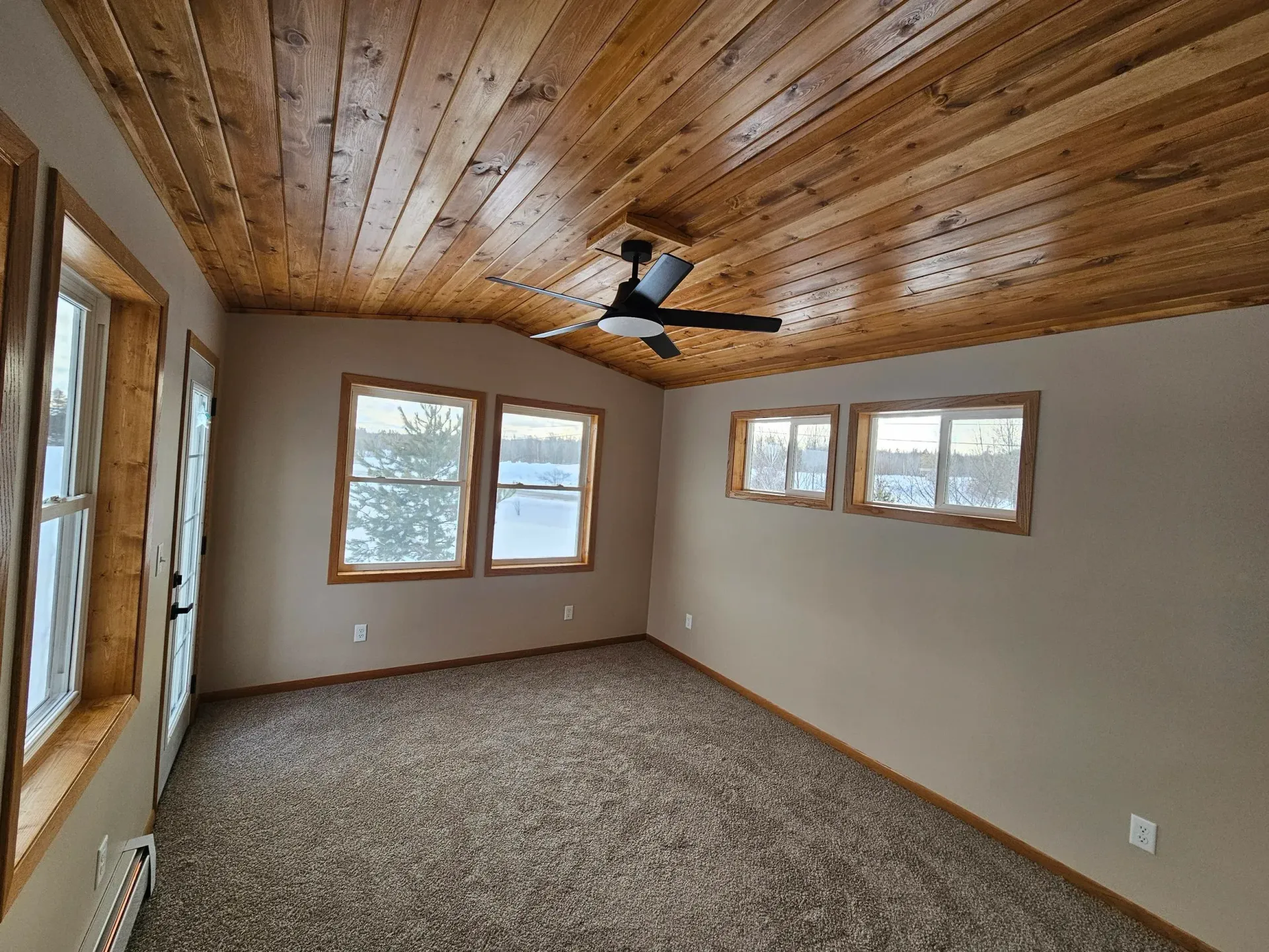 Empty room with wood-paneled ceiling, beige walls, carpeted floor, and several windows with a ceiling fan.