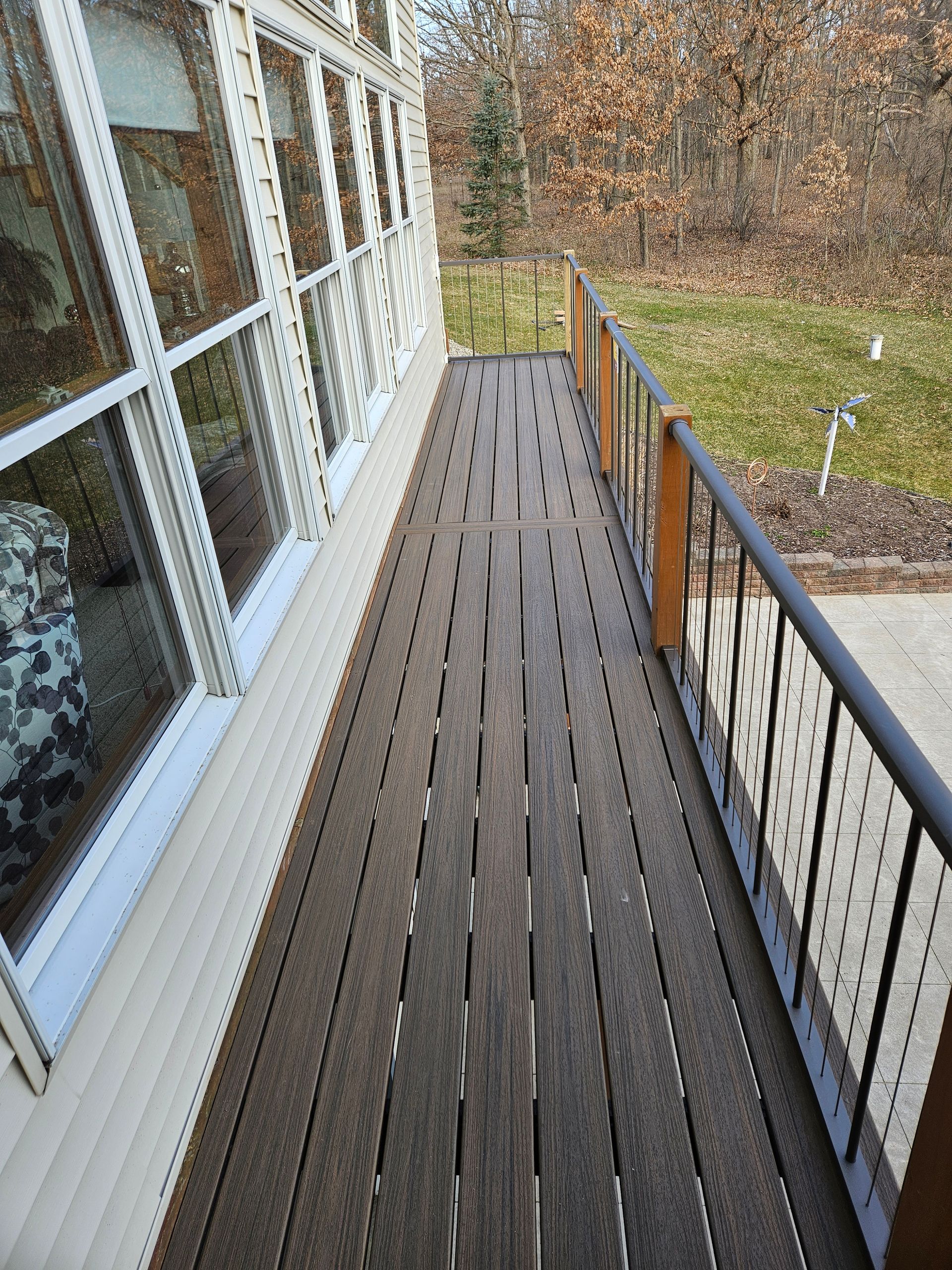 Narrow wooden balcony walkway beside a house, with railing and windows overlooking a yard.