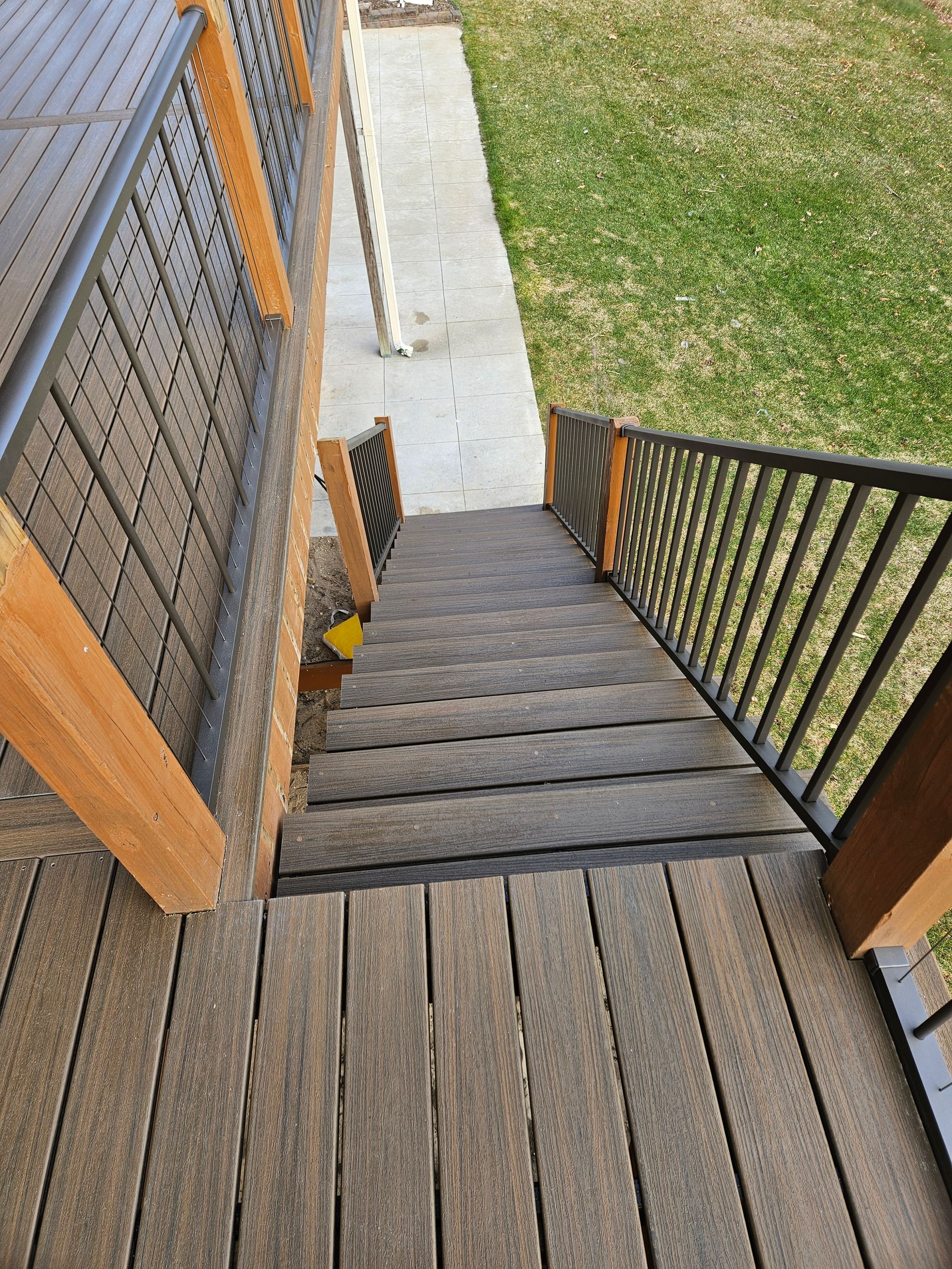 Wooden outdoor staircase descending to a concrete path beside a grassy yard and black railing.