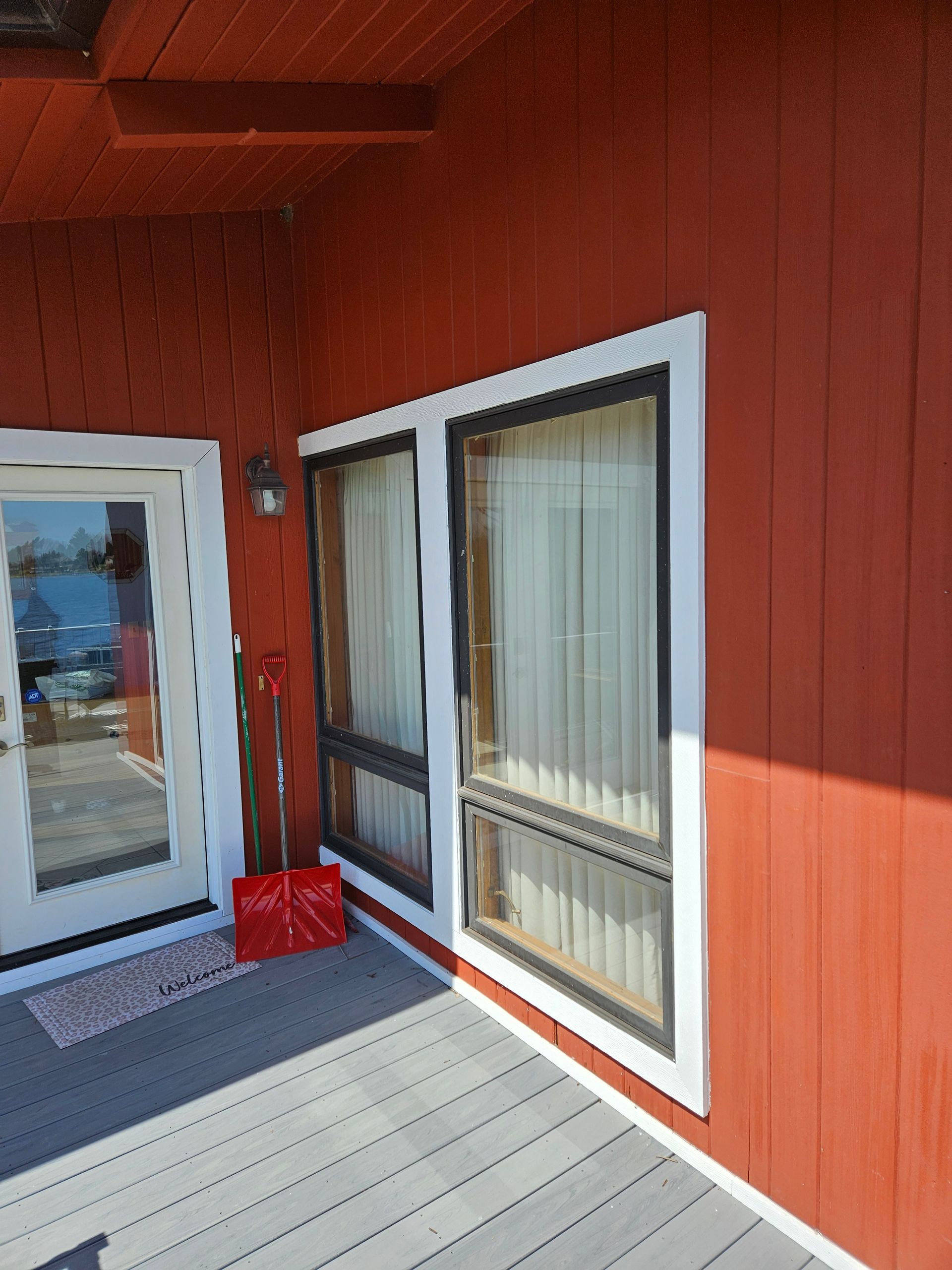Red wooden porch with white-trimmed door and window, sunlit deck, and a red broom by the entrance