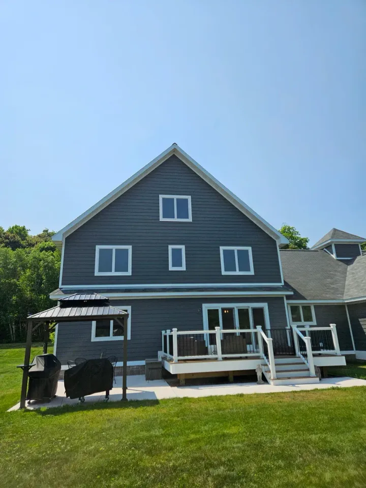 Dark gray house with white trim, deck, and grill on a green lawn under a blue sky