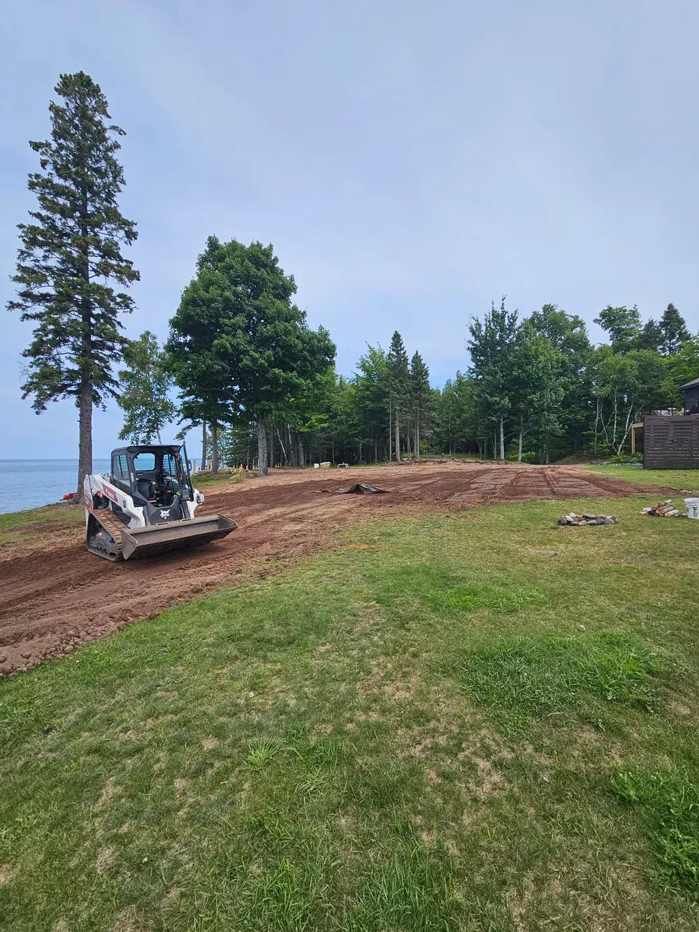 Bulldozer grading soil on a grassy lakeside lot with trees and a blue sky