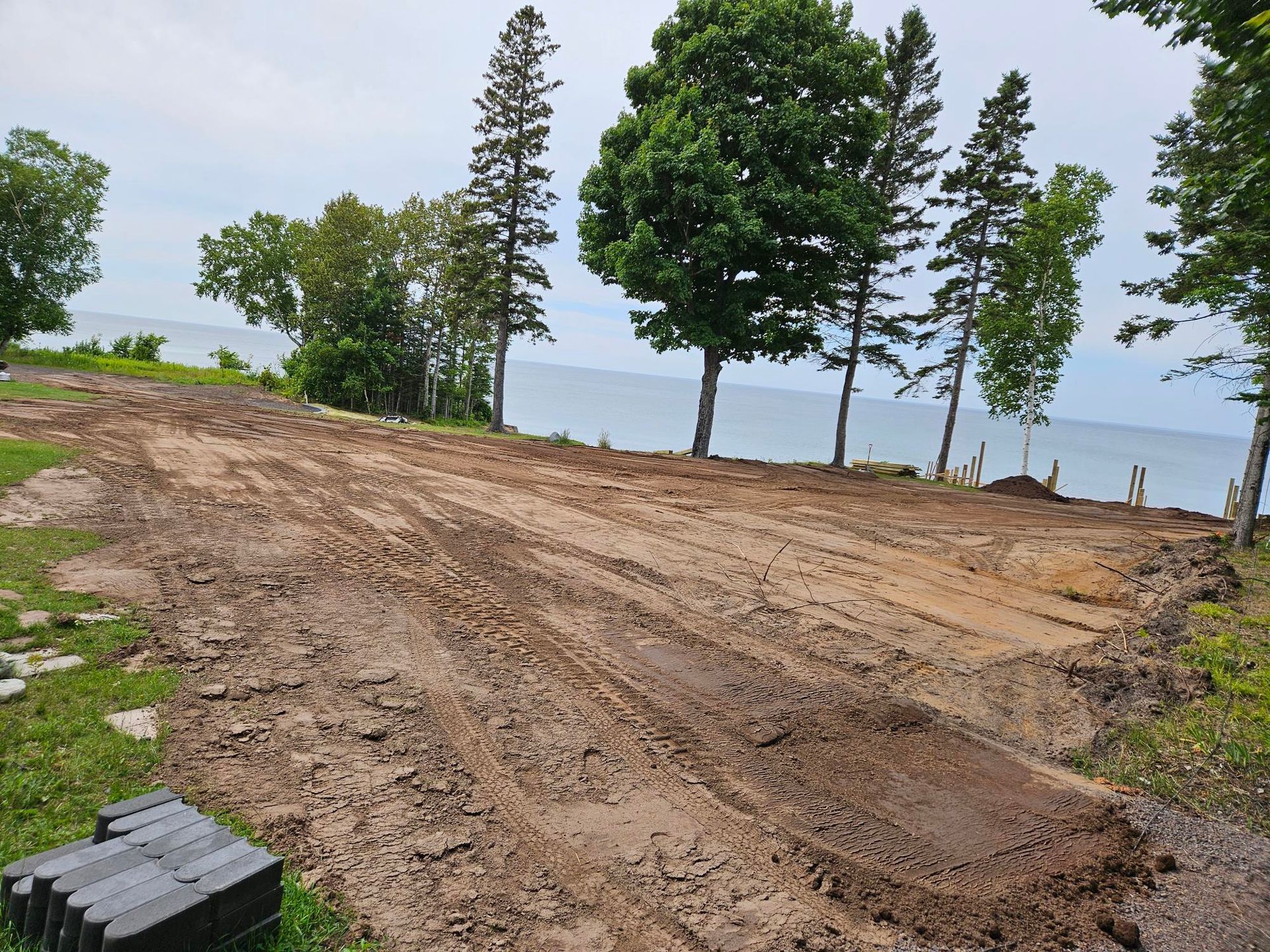 Cleared dirt lot by a lake with scattered trees and a cloudy sky