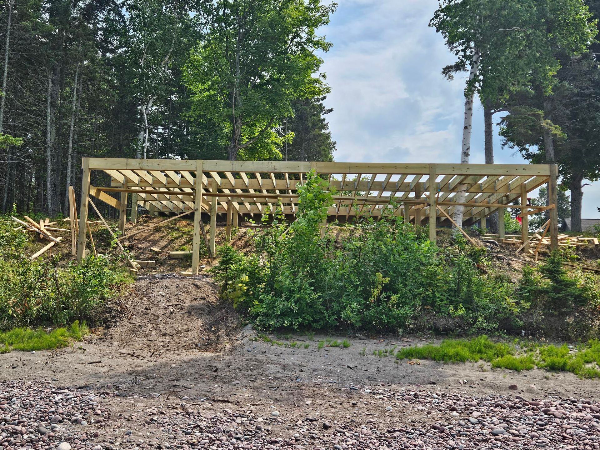 Wooden deck frame under construction on a muddy hillside surrounded by trees and greenery