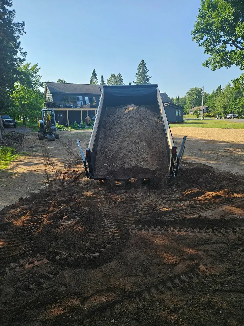Front loader dumping gravel in a muddy yard, with a house and trees in the background.