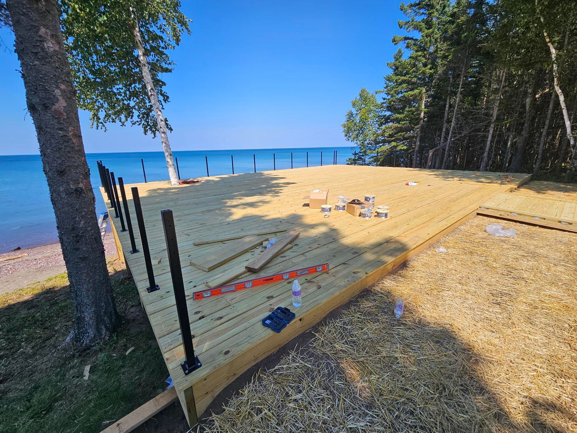 Beach volleyball court frame under construction on a sandy lakeshore, with trees and blue water in the background