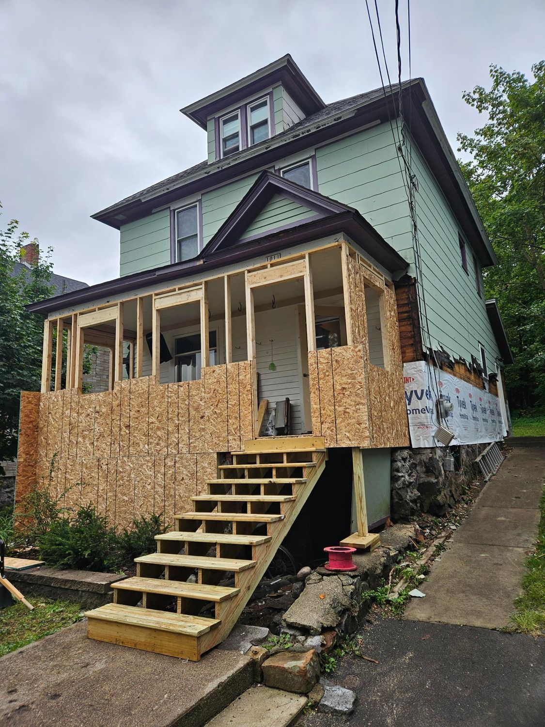 House under renovation with plywood-covered front porch and stairs along a sloped driveway