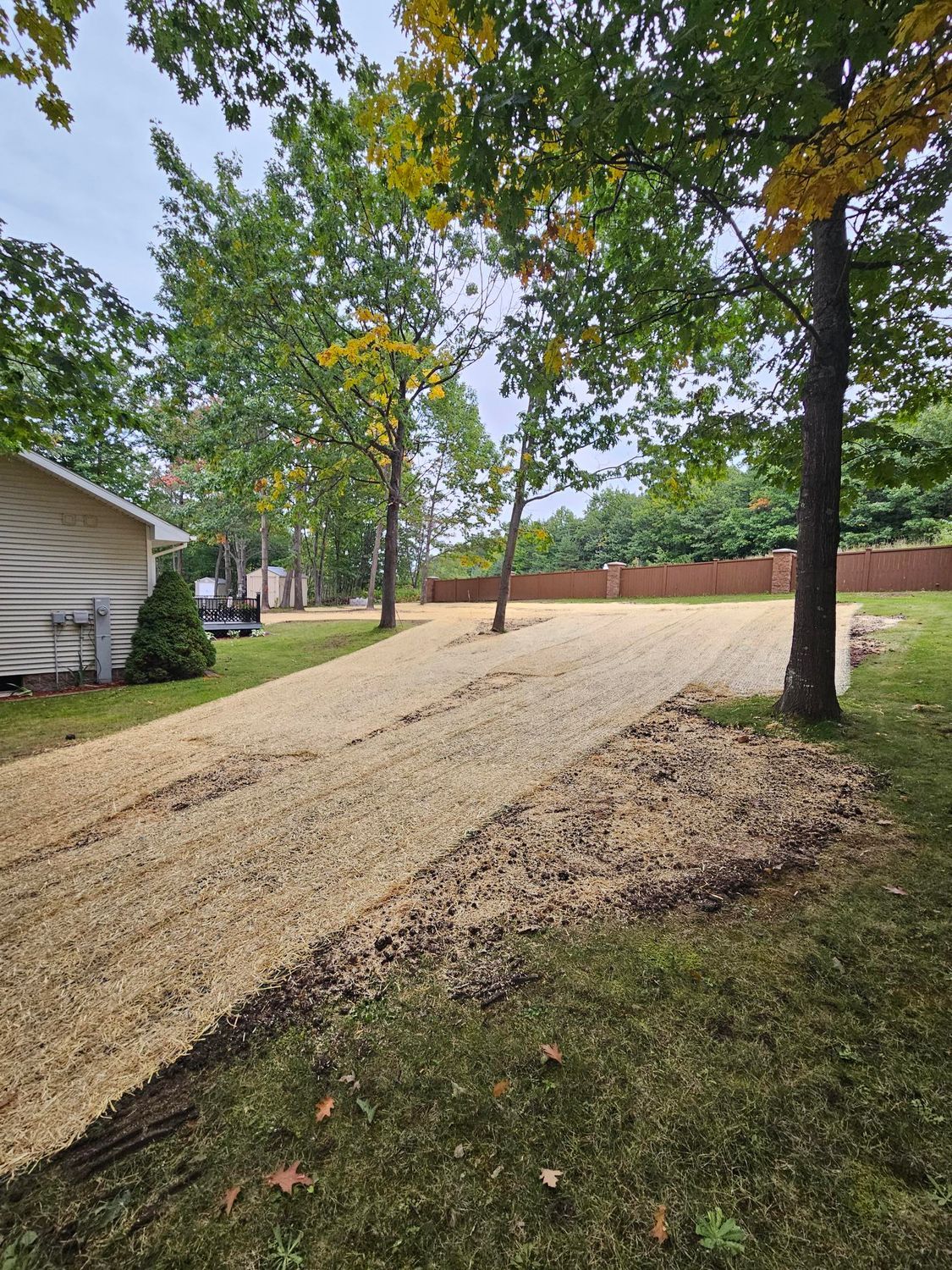 Gravel driveway beside a house and trees, with fallen autumn leaves and a brick wall in the background