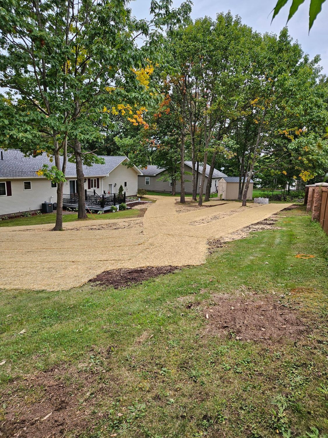 Gravel driveway beside houses and trees, with a grassy yard and overcast sky