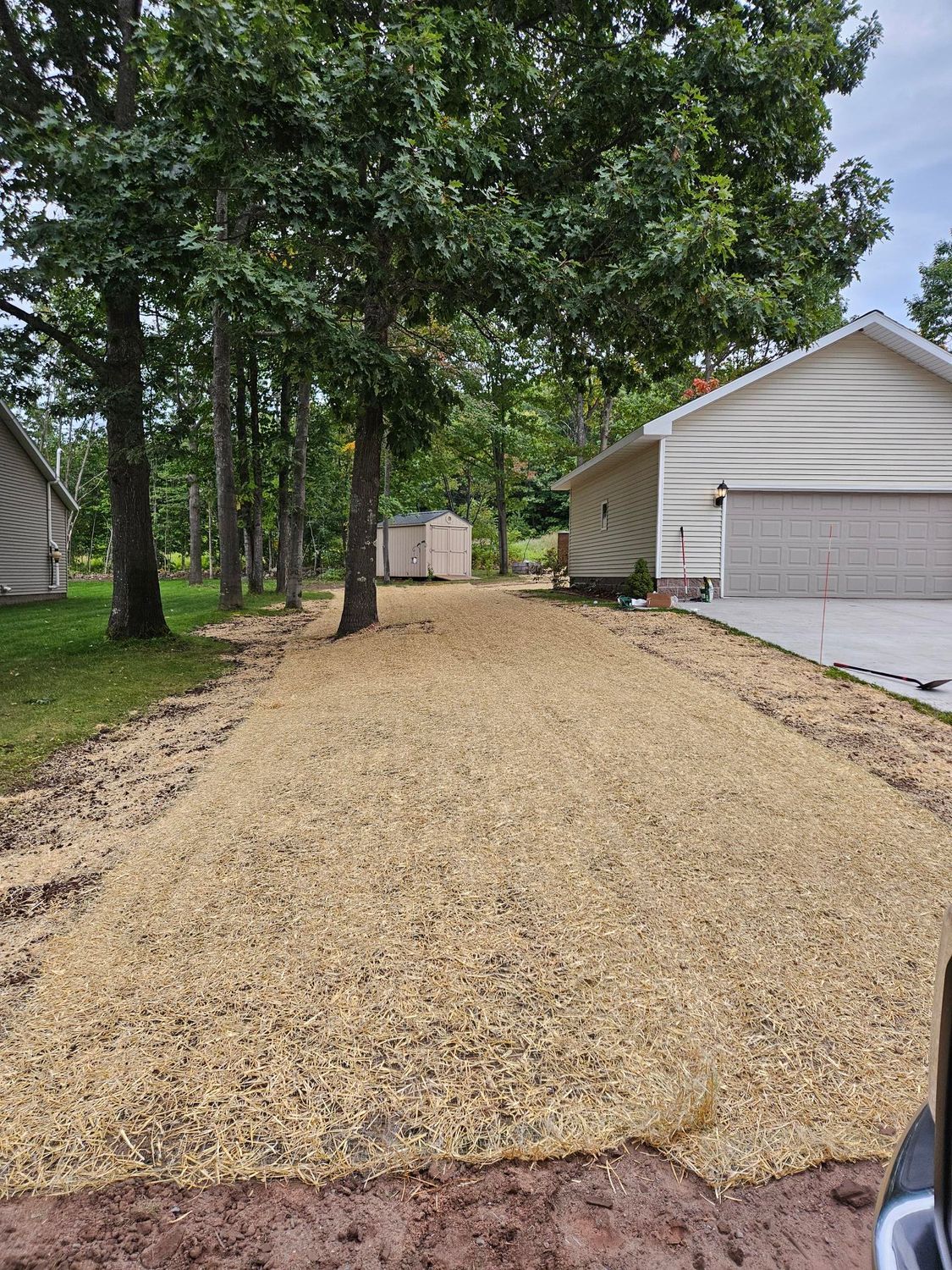 Fresh gravel driveway beside a house and wooded yard, with a garage and shed visible.