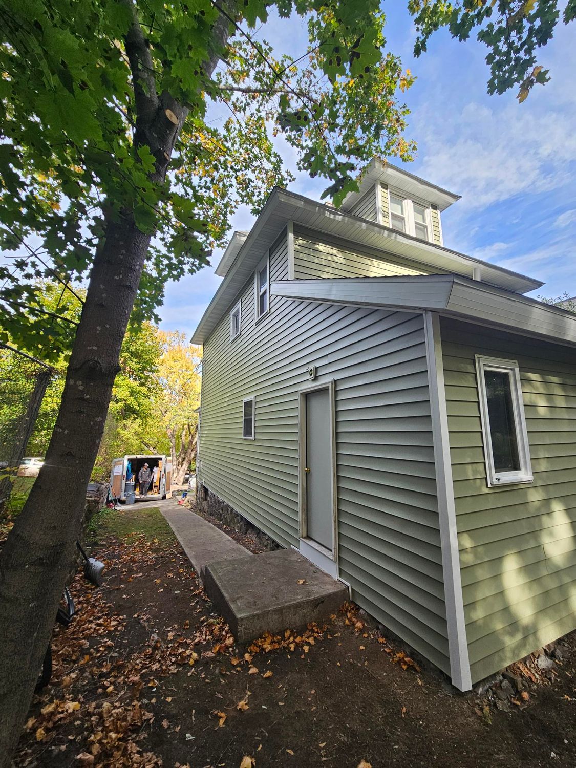 Green house side with white trim, trees overhead, and a narrow walkway along the foundation.