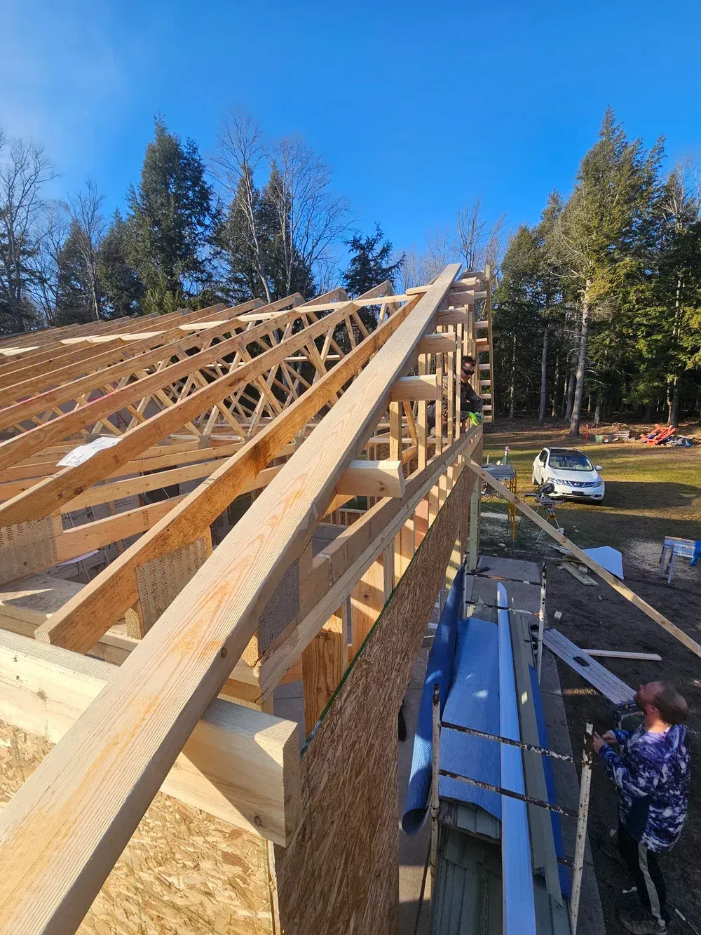 Roof framing under construction with wooden trusses and a worker below on a sunny day