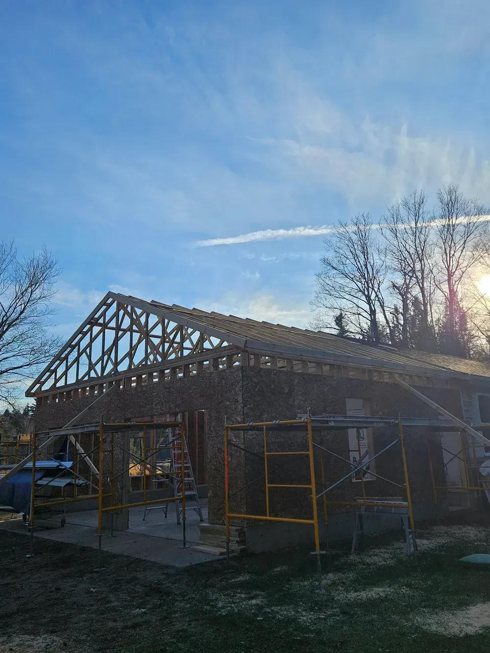 Partially built house frame with exposed roof trusses and scaffolding at sunset