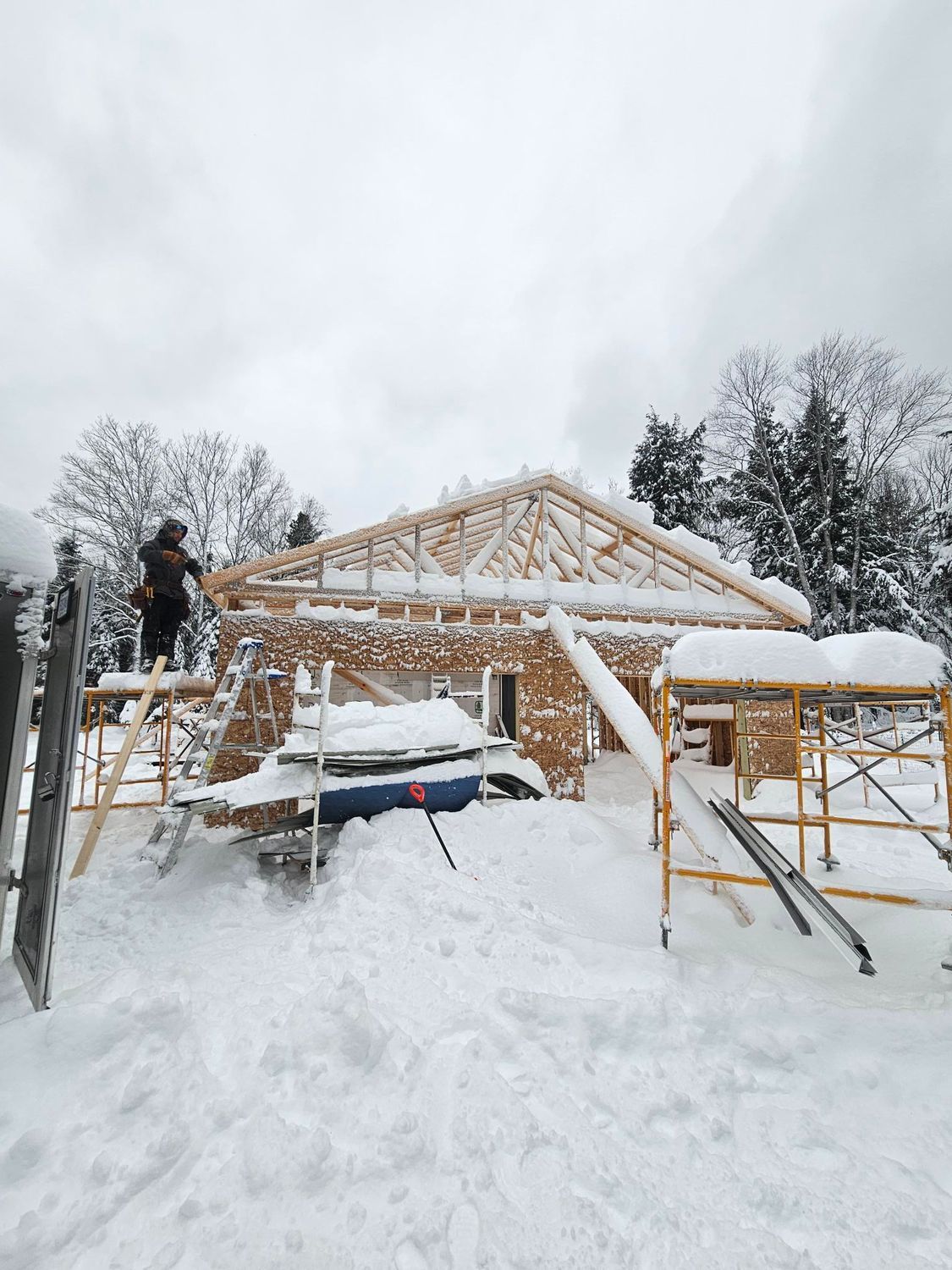 Snow-covered house under construction with wooden roof framing and a worker on a ladder.