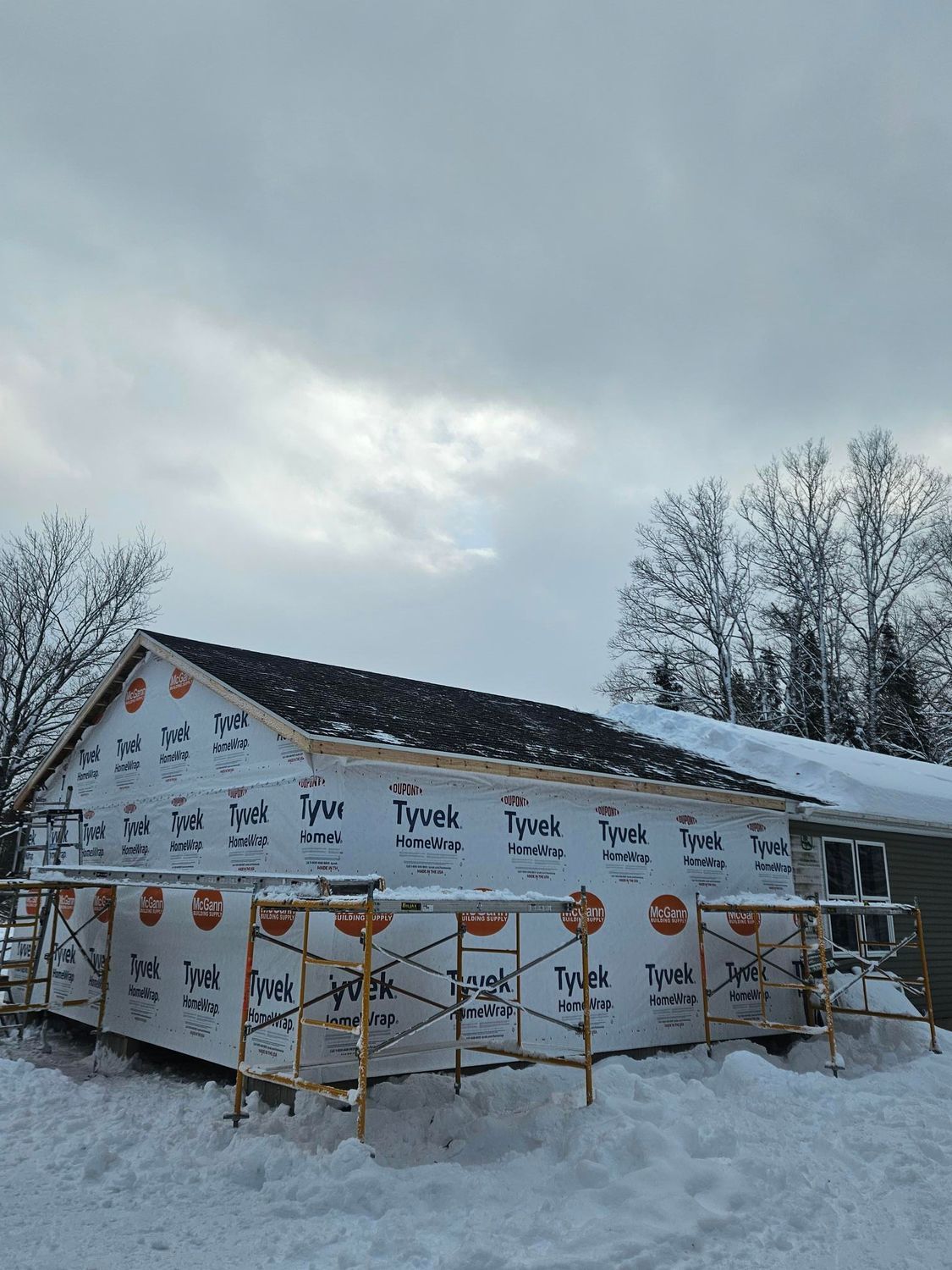 House under construction in snow, wrapped in Tyvek with scaffolding and bare trees nearby