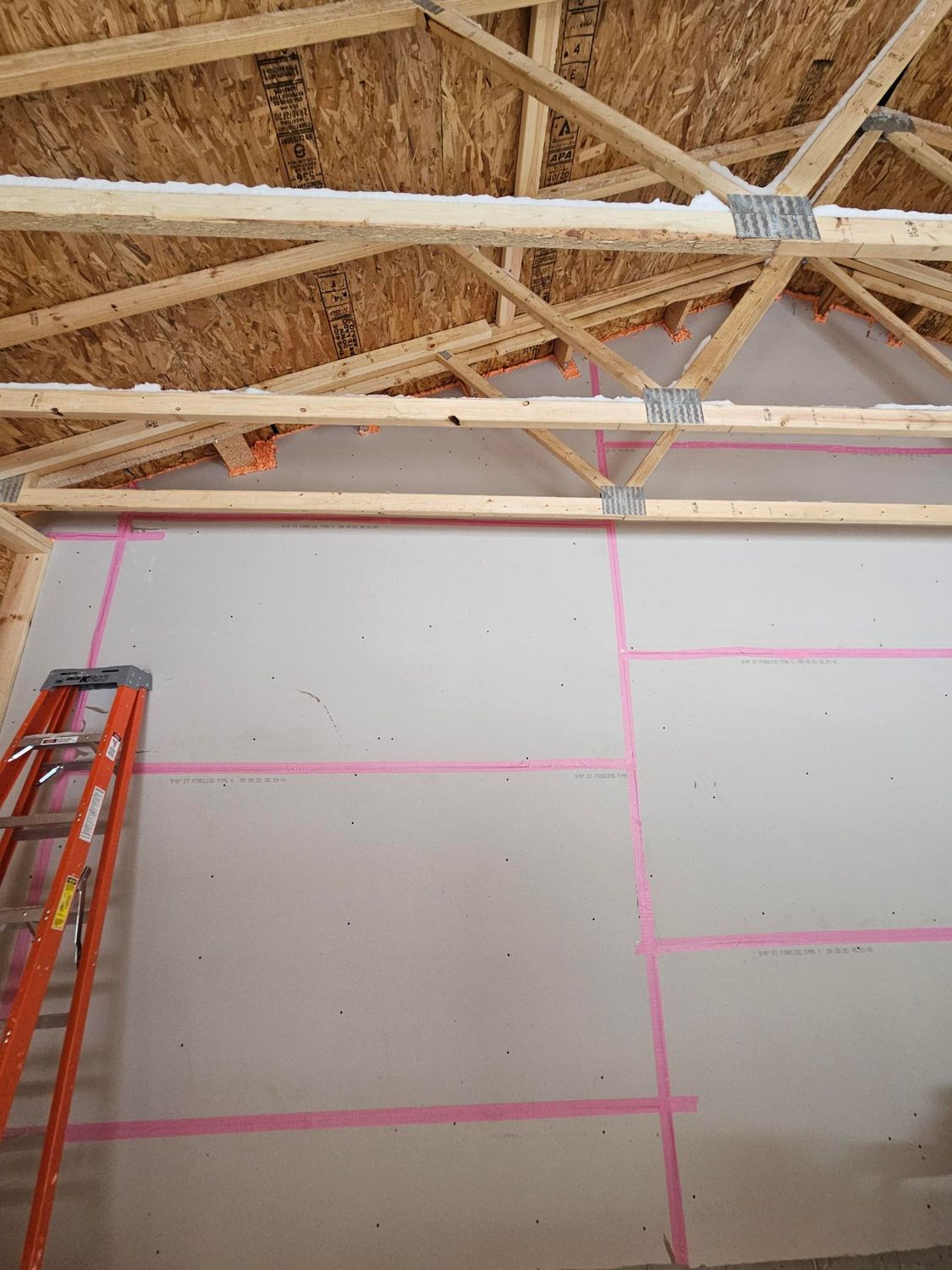 Interior wall under construction with exposed roof trusses, drywall panels, and a red ladder on the left.