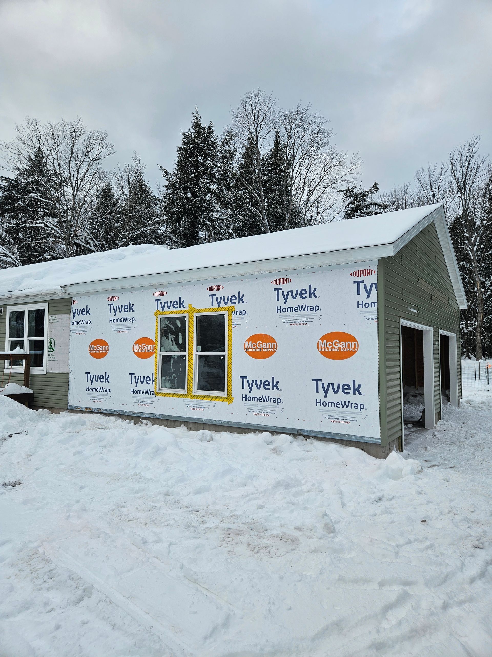 Snowy construction site with a partially built house wrapped in Tyvek and yellow-framed windows.