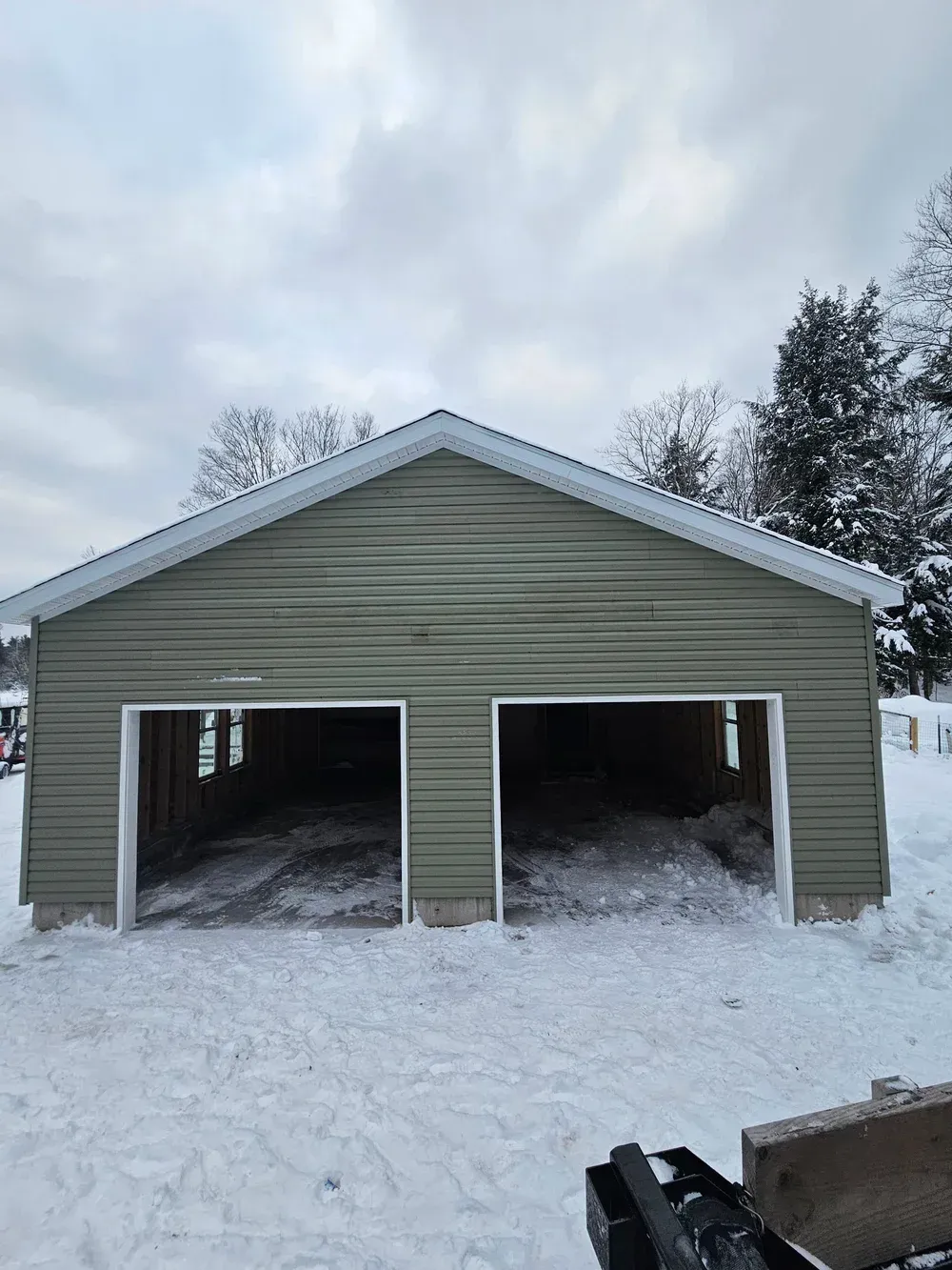Snow-covered two-bay garage with green siding and open doors under a cloudy winter sky