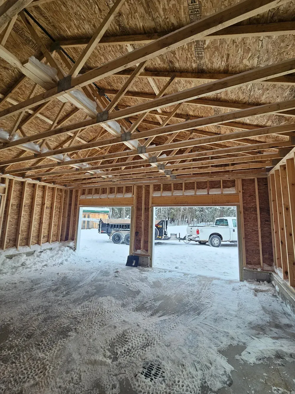 Unfinished garage interior with exposed wooden trusses and snow on the floor, opening to parked vehicles outside