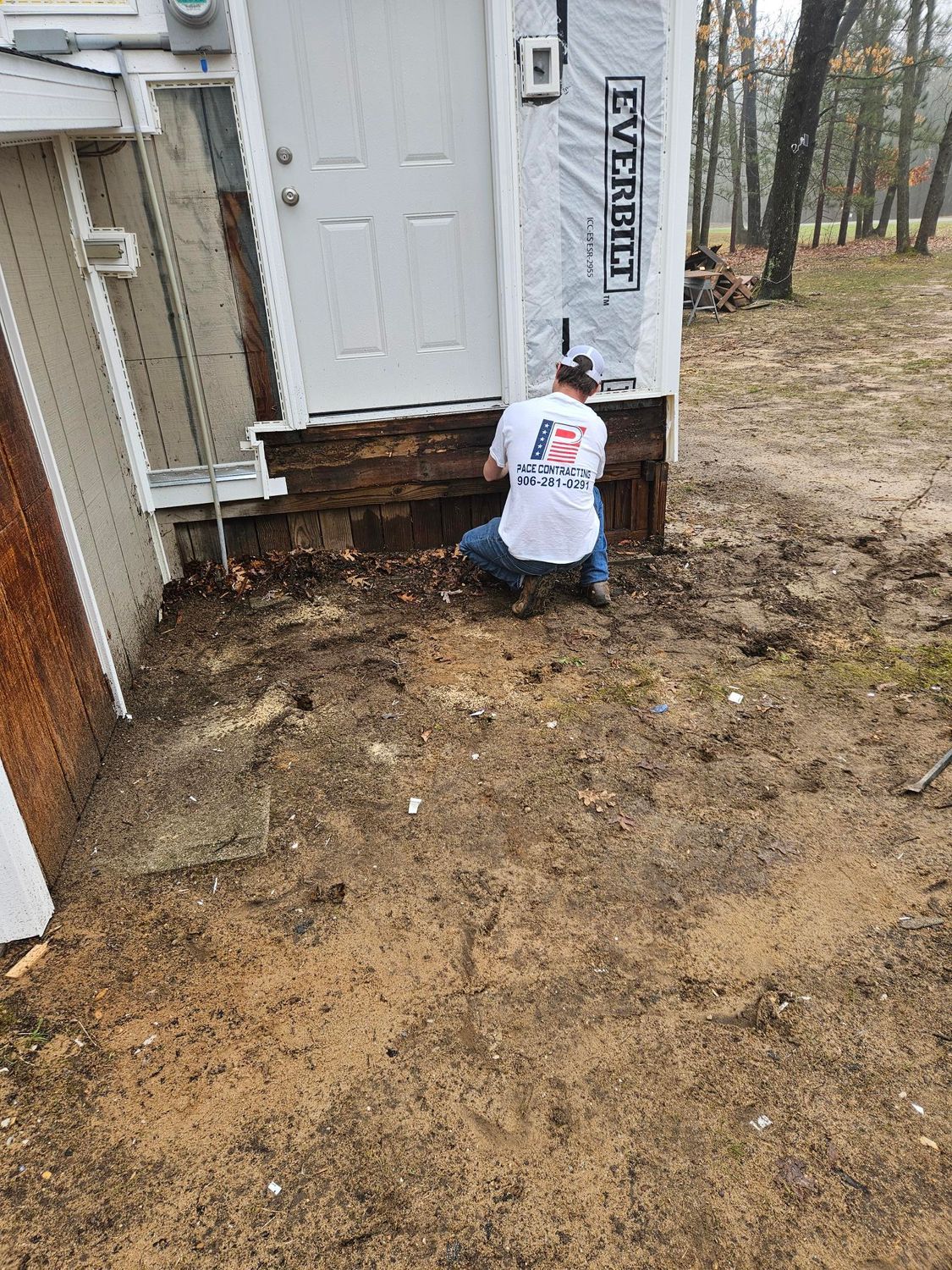 Worker repairs wet soil near house entry beside a white door and downspout.