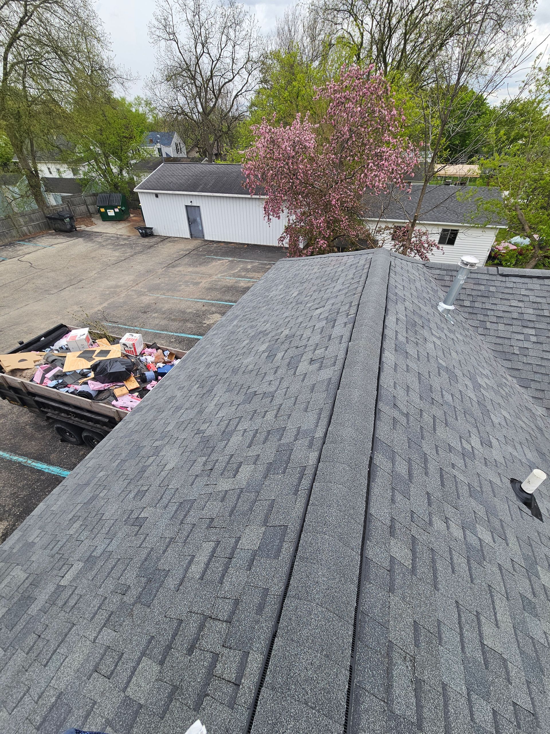 Rooftop view of gray shingles with a tree and garage in the background, and trash bins at left.