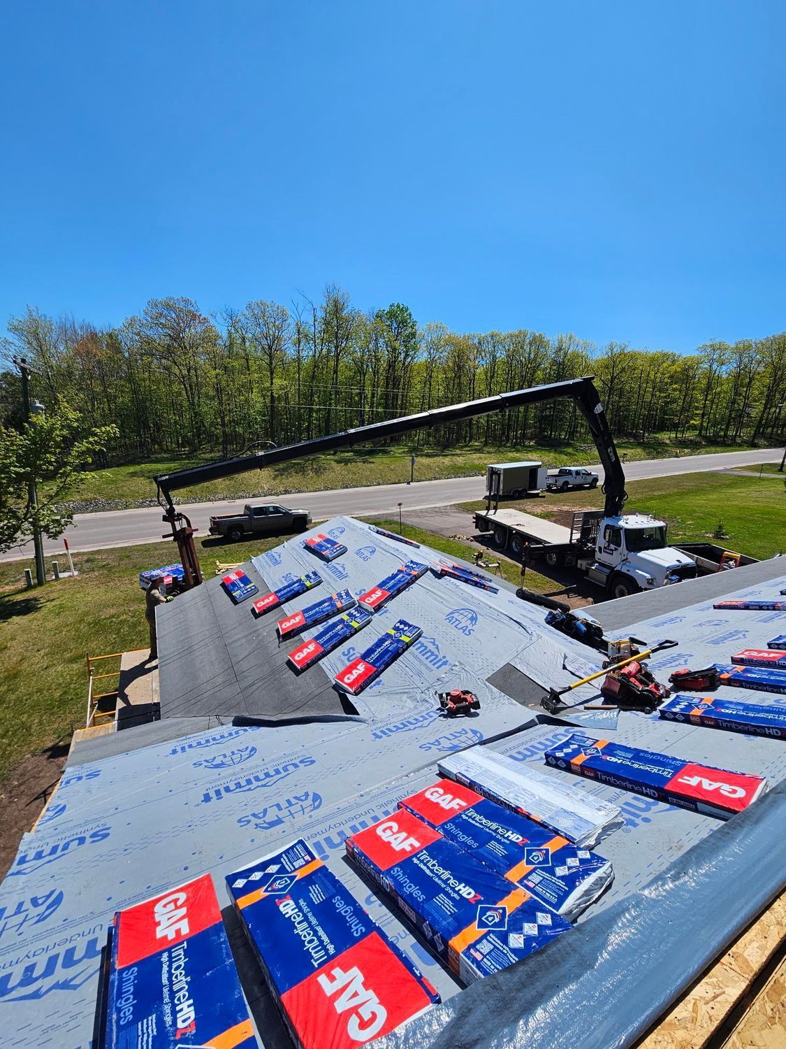 Roof under construction with blue underlayment and a crane arm over a sunny hillside site