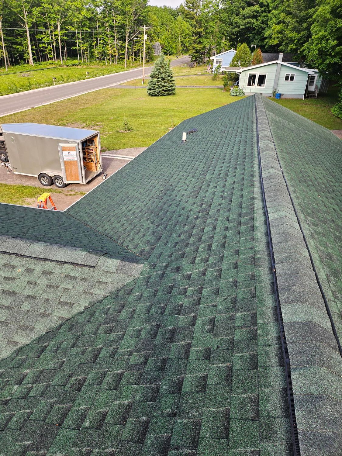 View from a green shingle roof toward a grassy yard, trees, a trailer, and a small white building