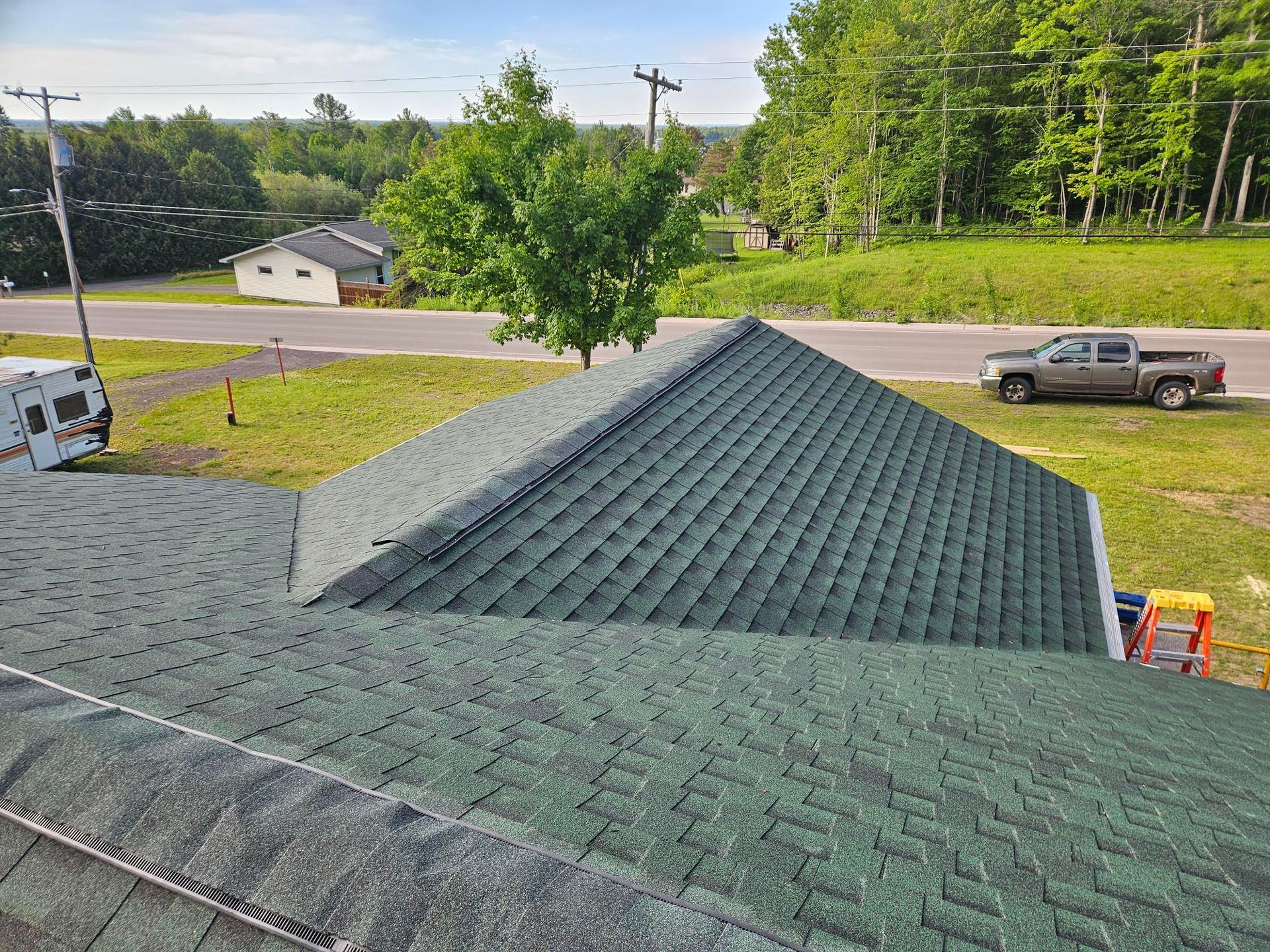 View from a roof overlooking a green shingle roof, road, parked truck, and trees on a sunny day