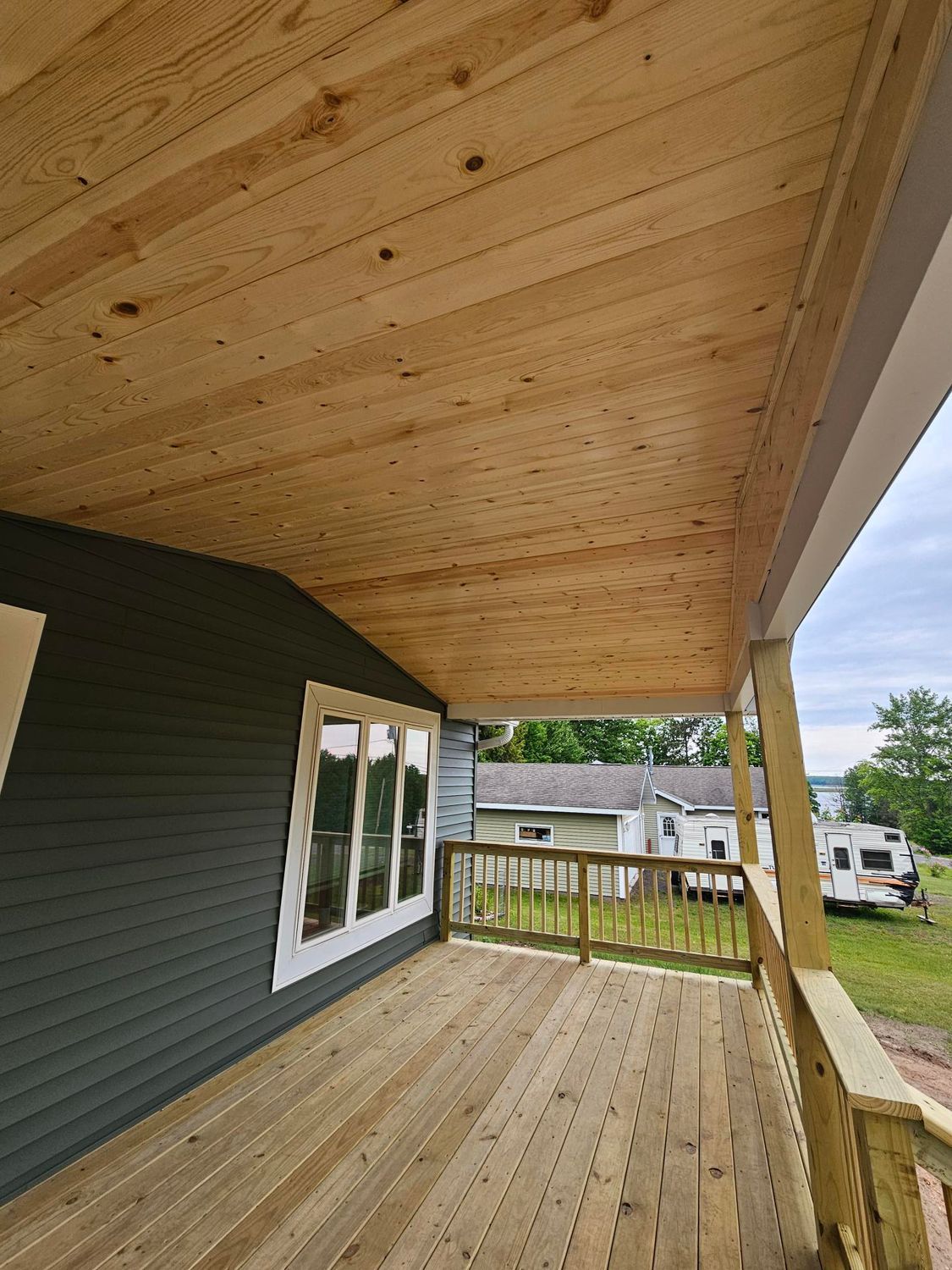 Covered wooden porch with a pine ceiling, green siding, railing, and a view of a yard and parked RVs.