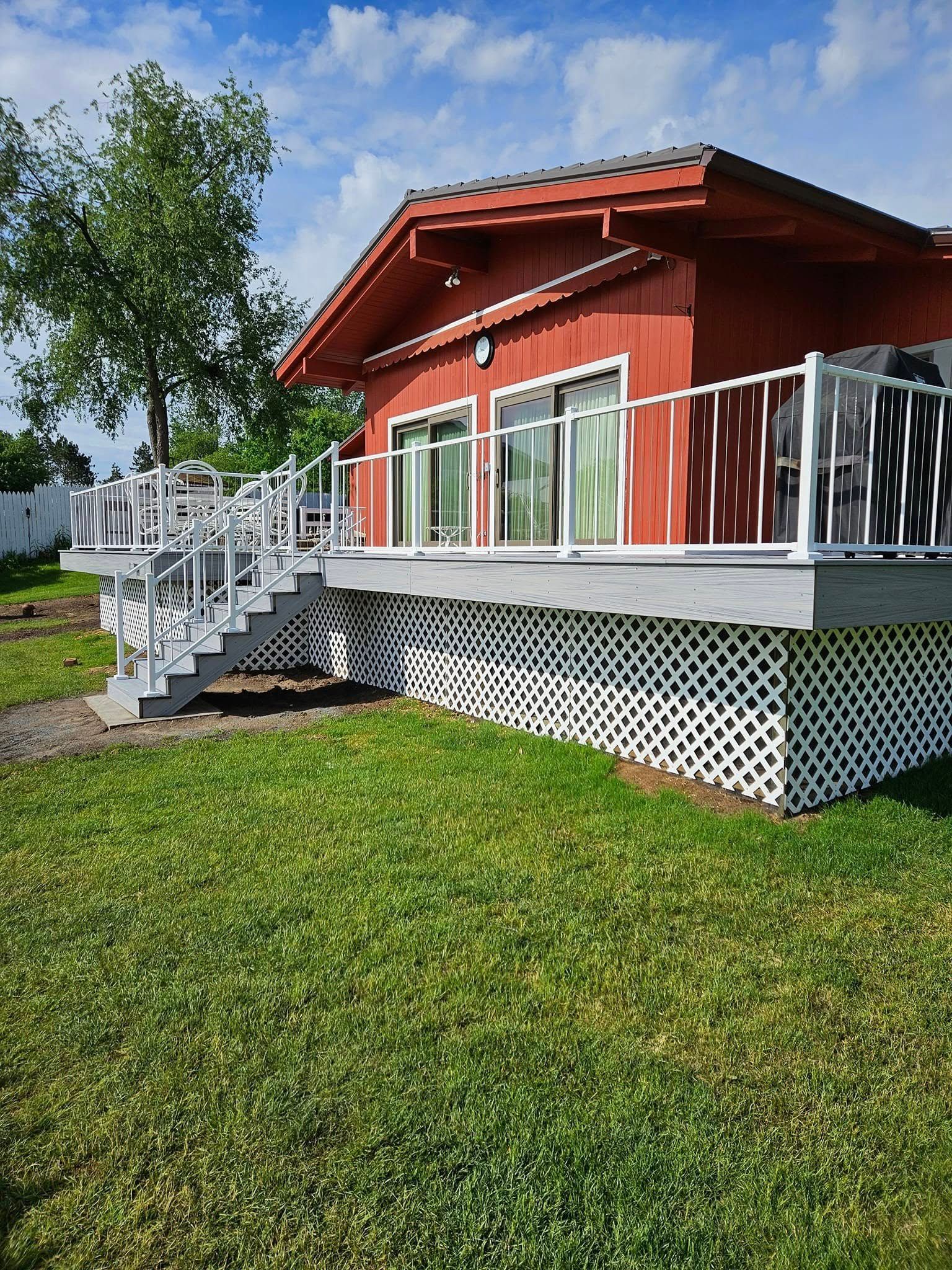Red house with white deck railing and stairs, set on a green lawn under a tree
