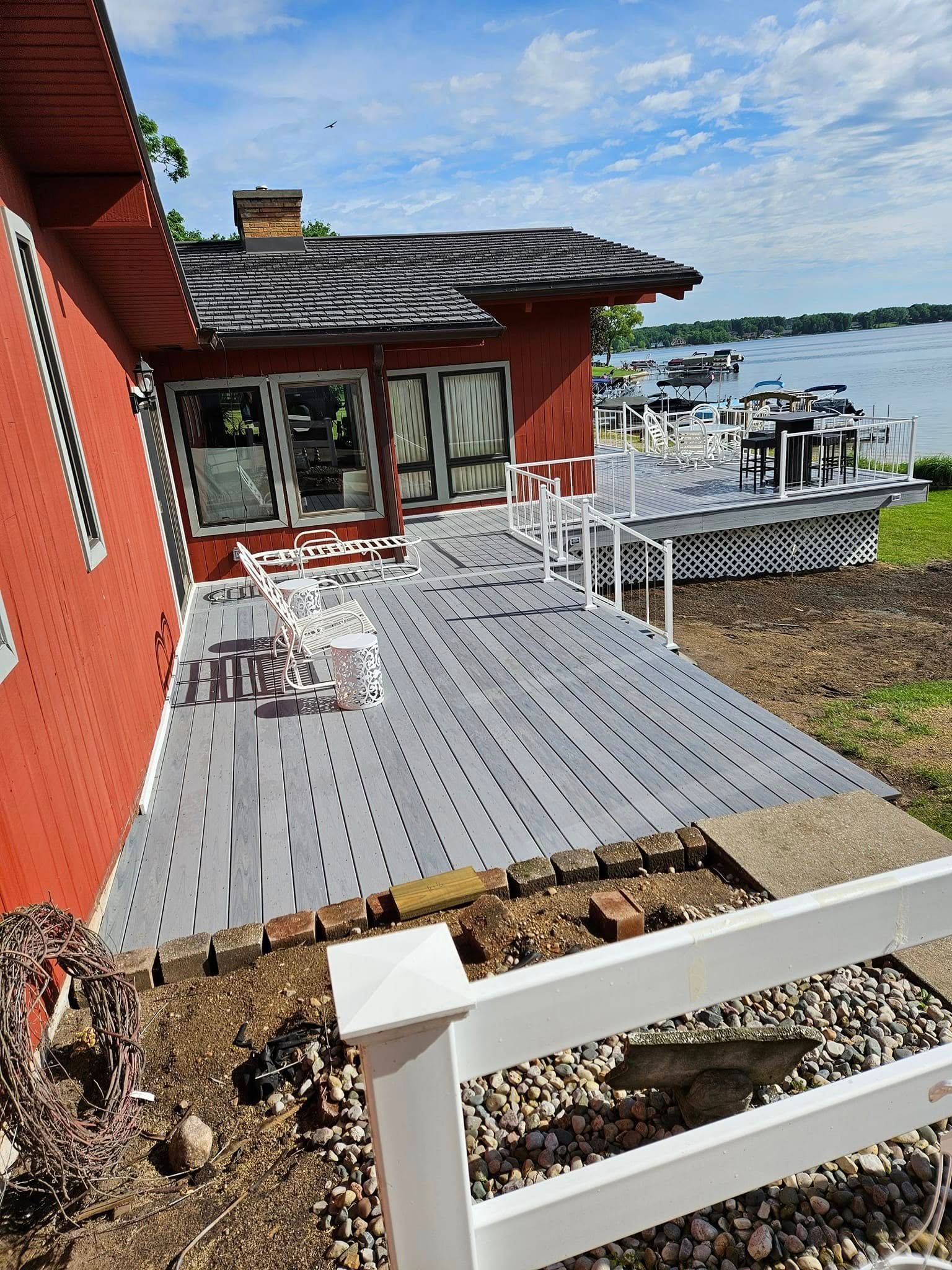 Red lakeside house with a gray deck, white railing, and patio furniture under a blue sky