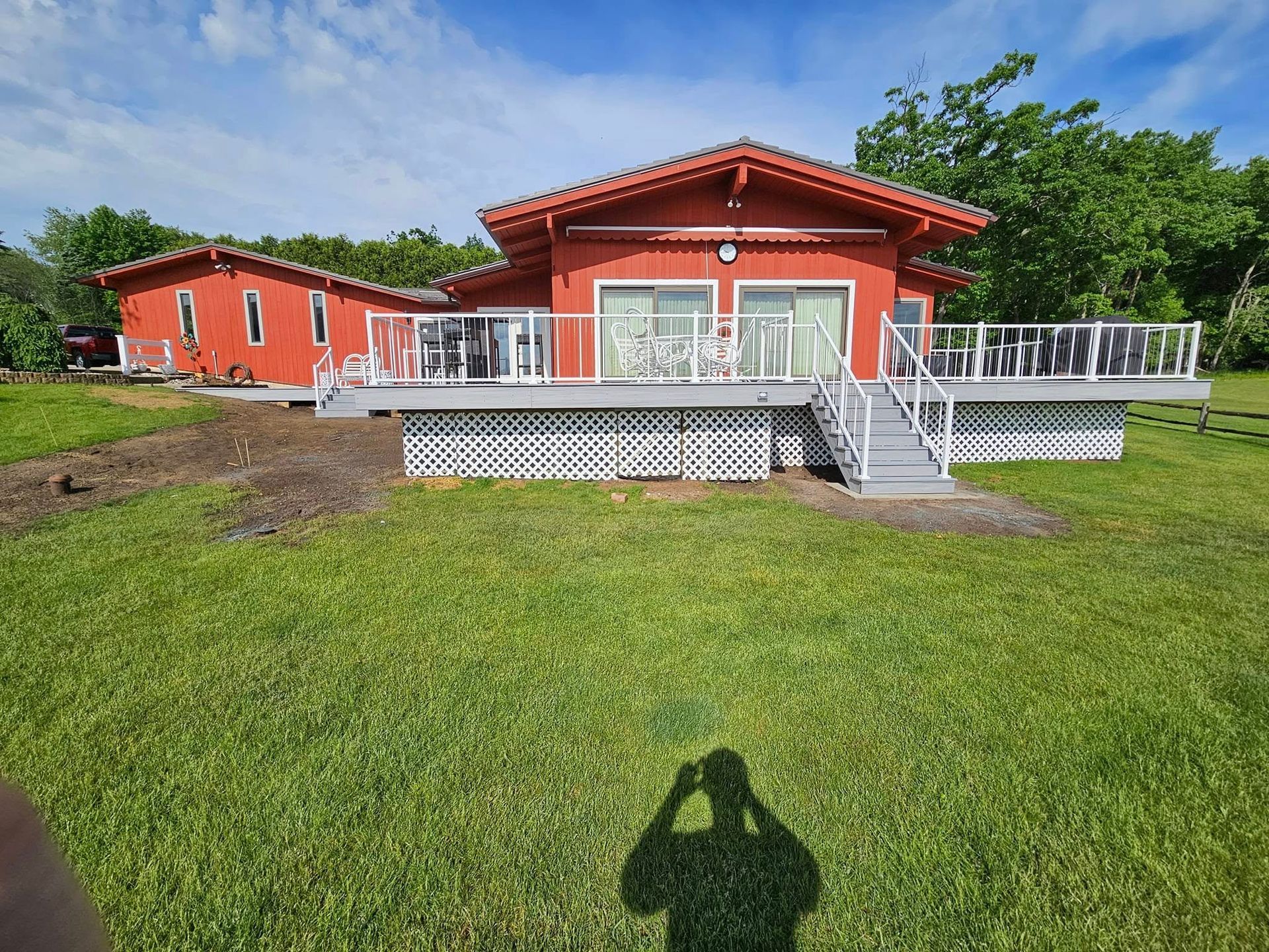 Red house with white deck and stairs on a grassy lawn, surrounded by trees.