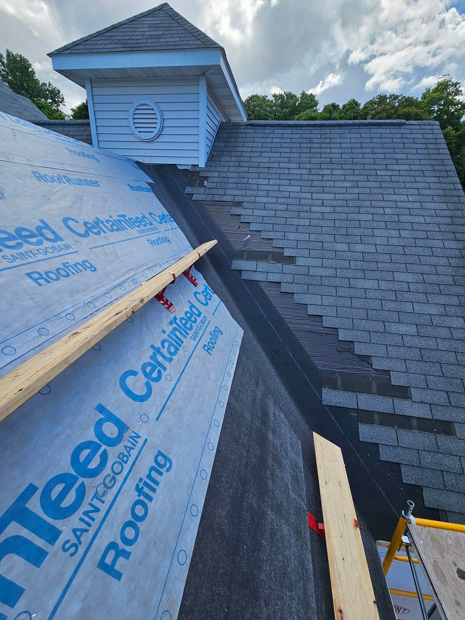 Roof under construction with shingled section, blue underlayment, and dormer vent against a cloudy sky
