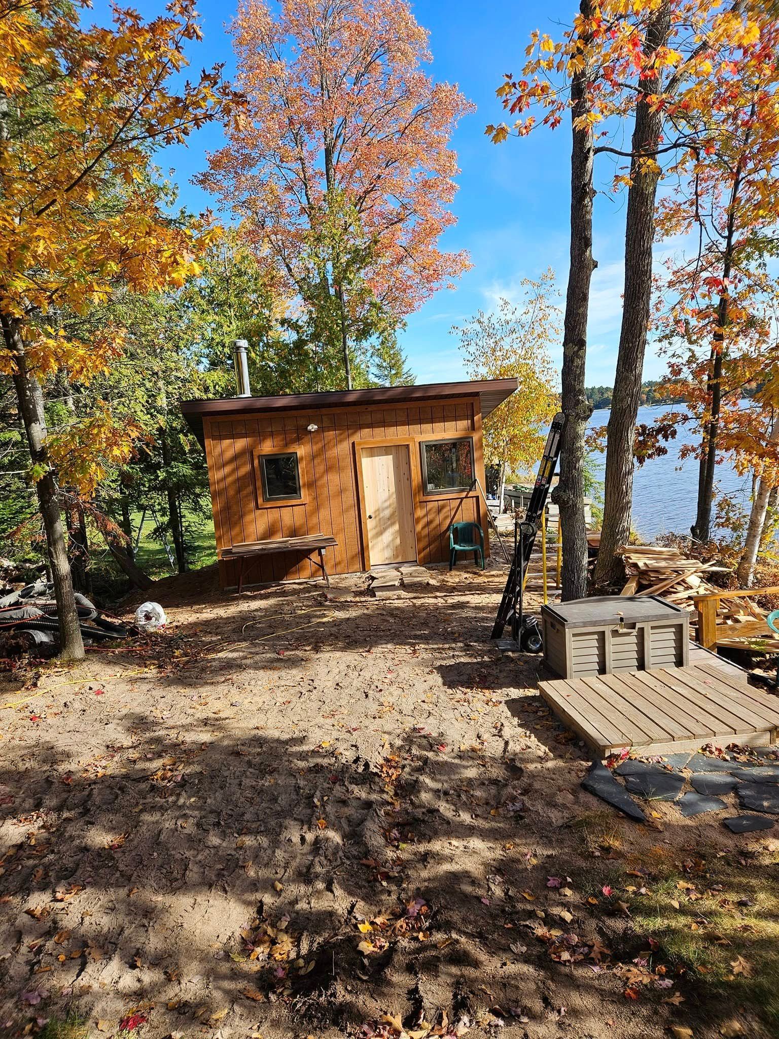 Small wooden cabin in an autumn forest with lake view, surrounded by colorful trees and stacked firewood.