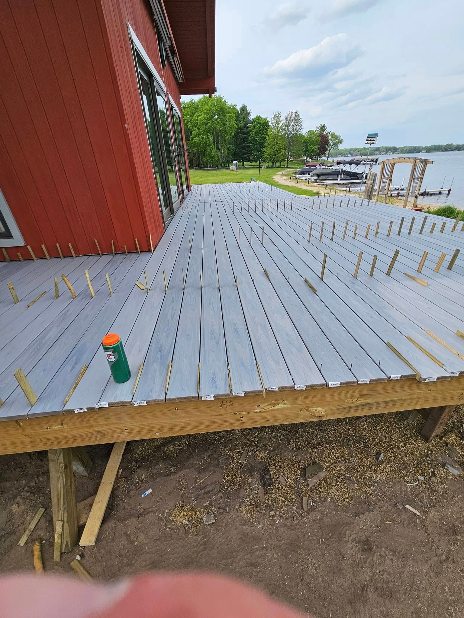 Gray deck under construction beside a red house, with exposed beams and a green bottle on the floor.