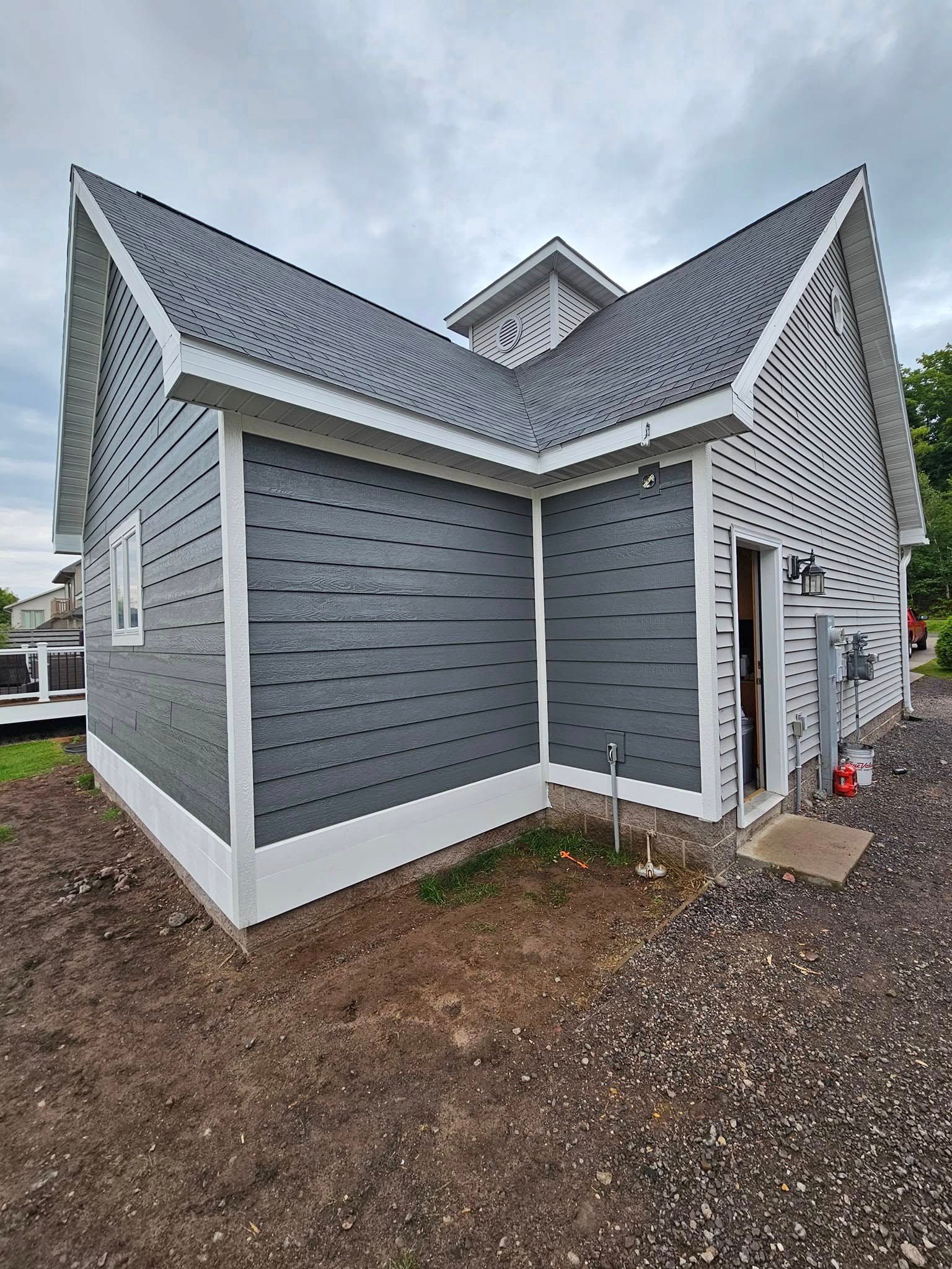 Gray house exterior with steep gabled roof, white trim, and gravel lot under cloudy sky