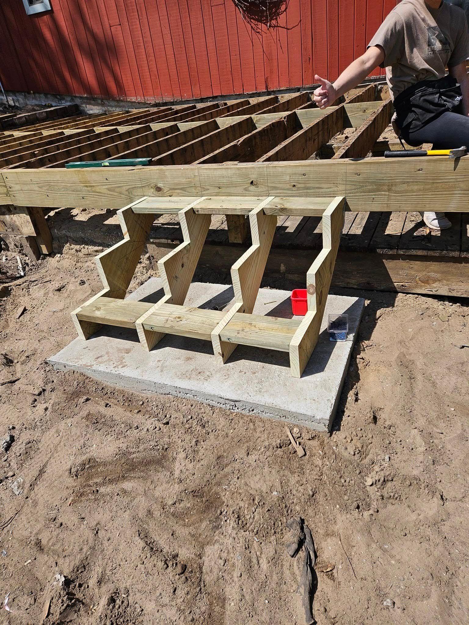 Person building a wooden bench frame on a concrete base in a dirt yard, with a red tool nearby
