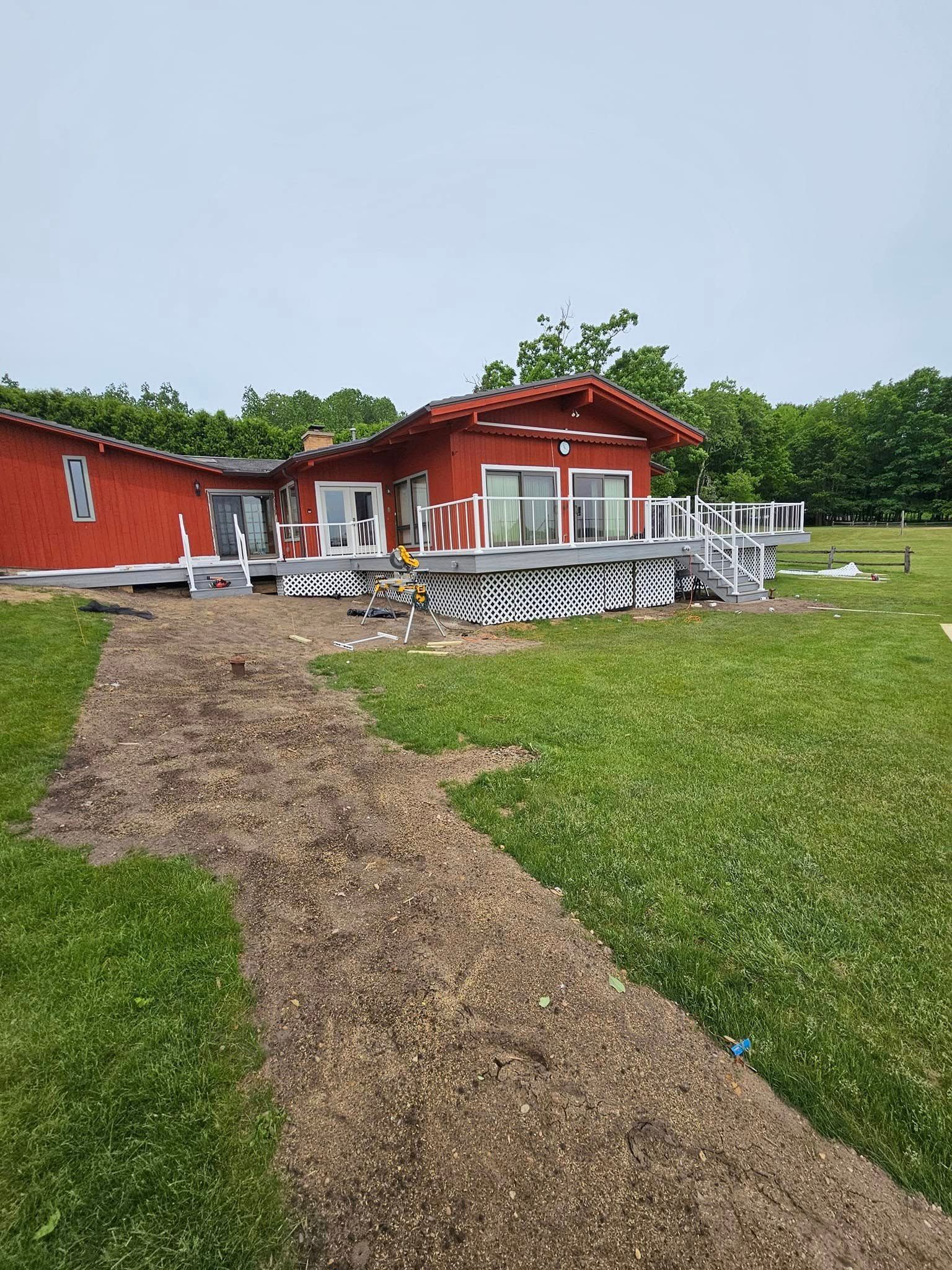 Red house with white deck and chairs beside a grassy path under a cloudy sky.