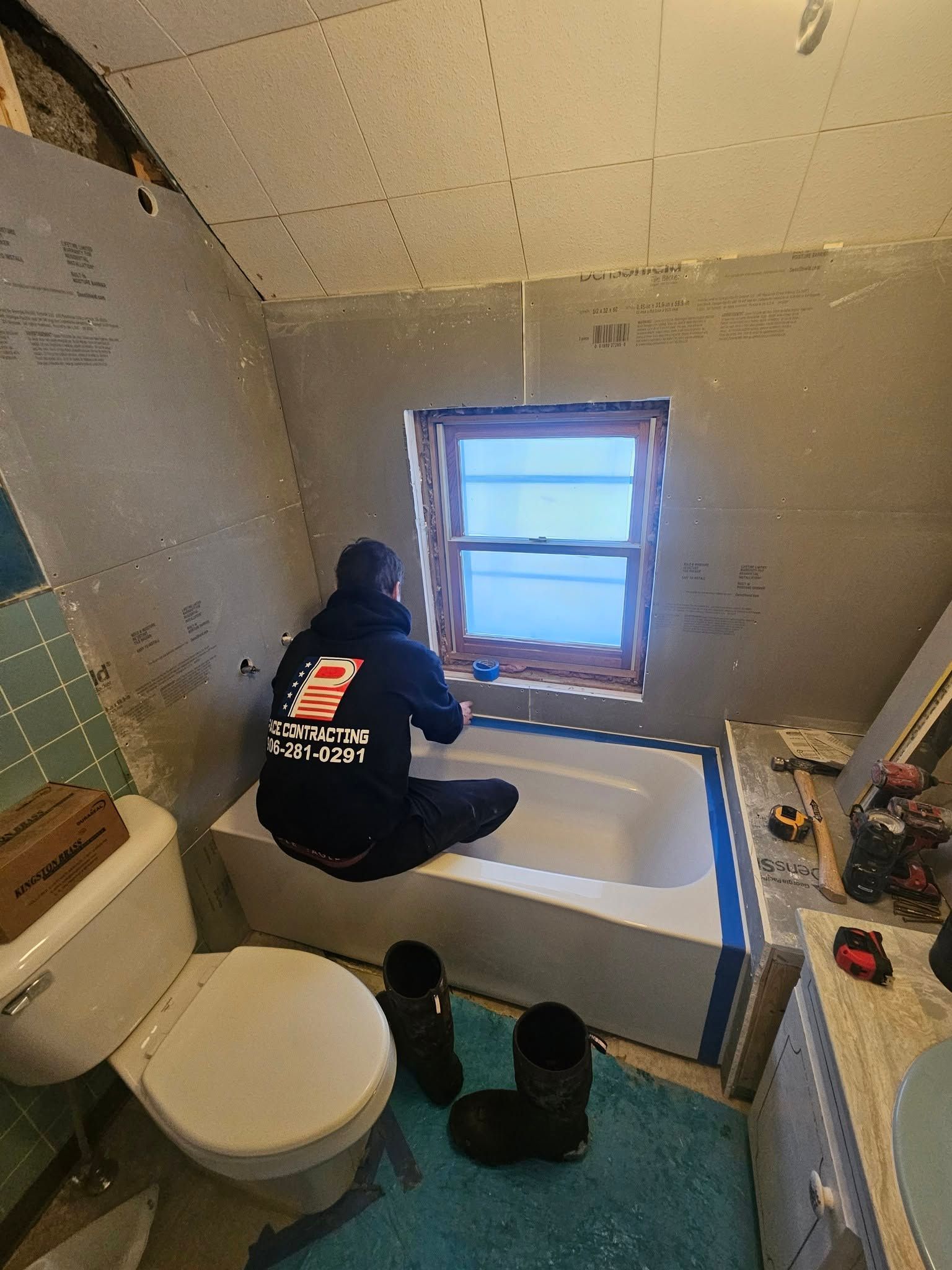 Person kneels by a bathroom window, working on a white tub or shower surround during renovation.