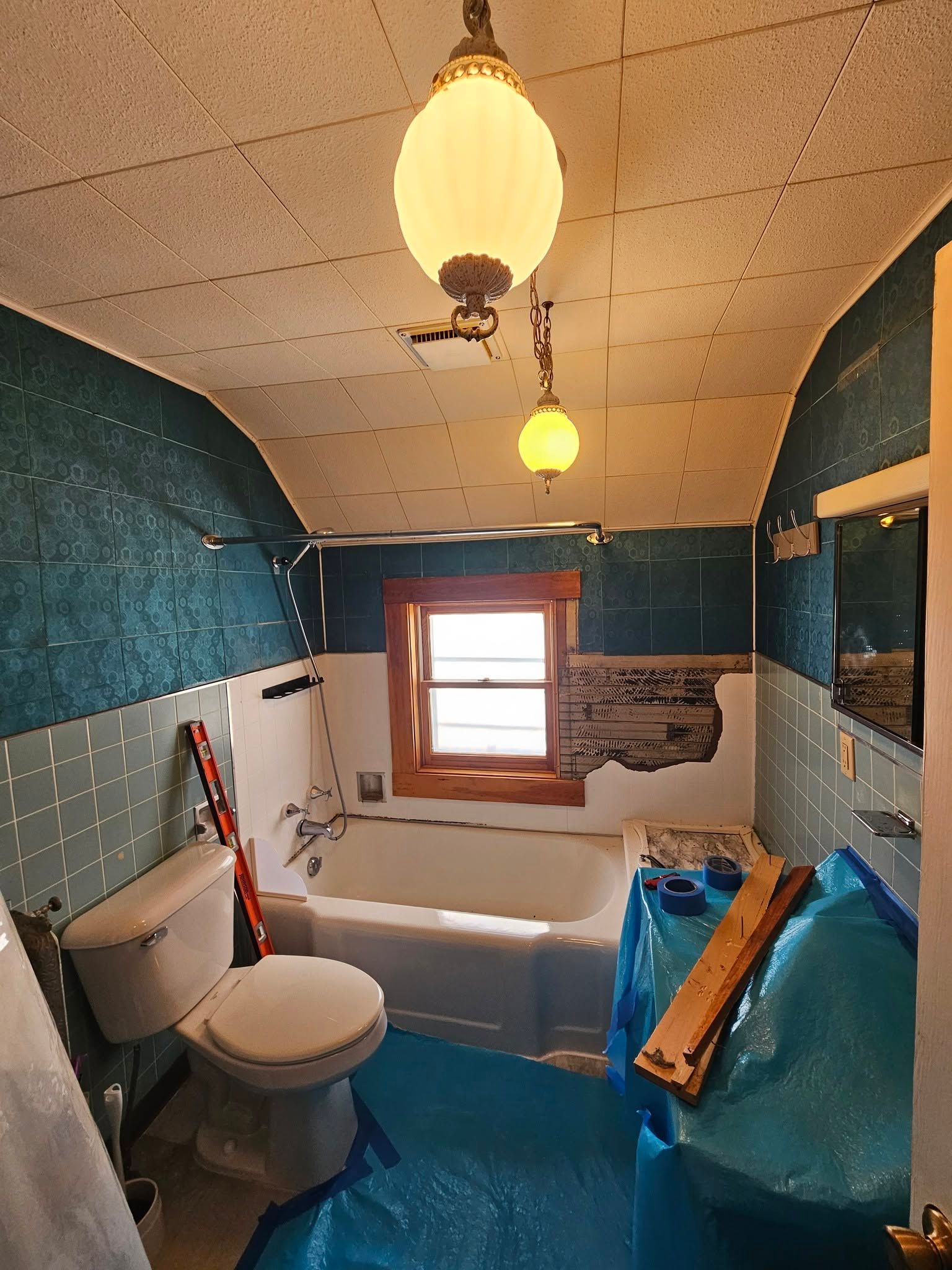 Small bathroom under renovation with a toilet, bathtub, window, and blue protective coverings.