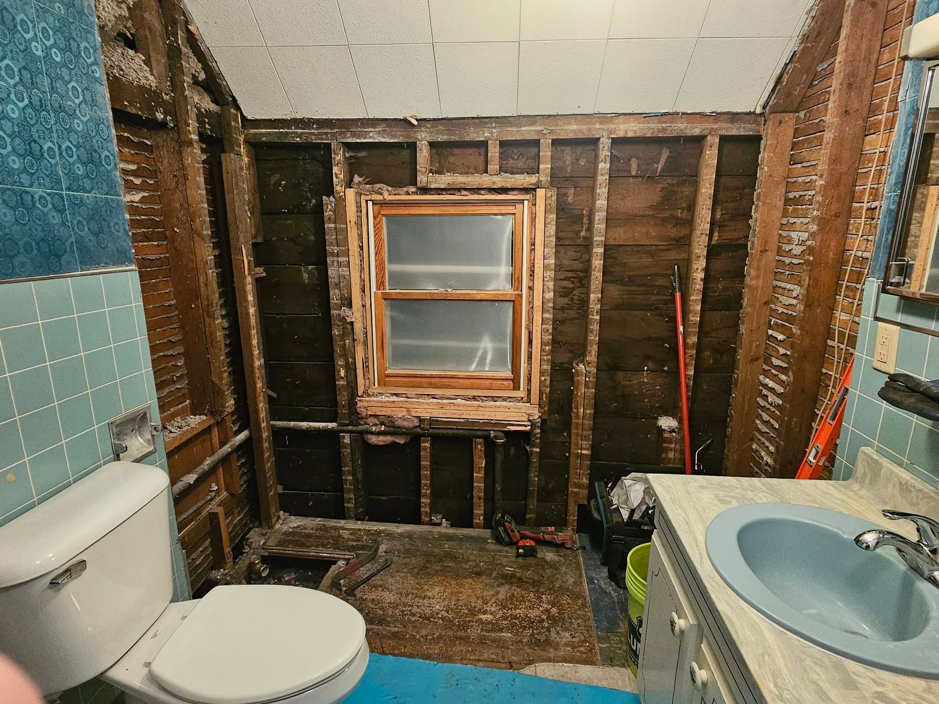 Partially gutted bathroom with exposed studs, toilet, sink vanity, and a small window above the back wall